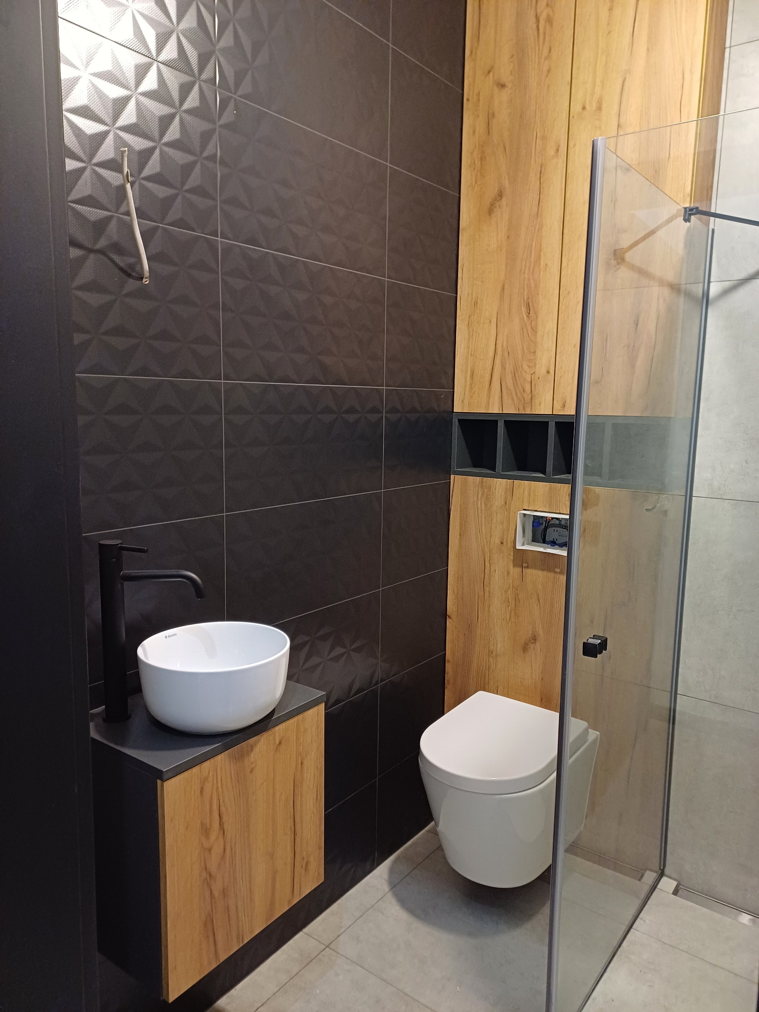 Nowoczesna łazienka z czarnymi, trójwymiarowymi płytkami, drewnianymi szafkami i białą ceramiką. Uwagę zwraca minimalistyczna umywalka nablatowa i kabina prysznicowa typu walk-in.