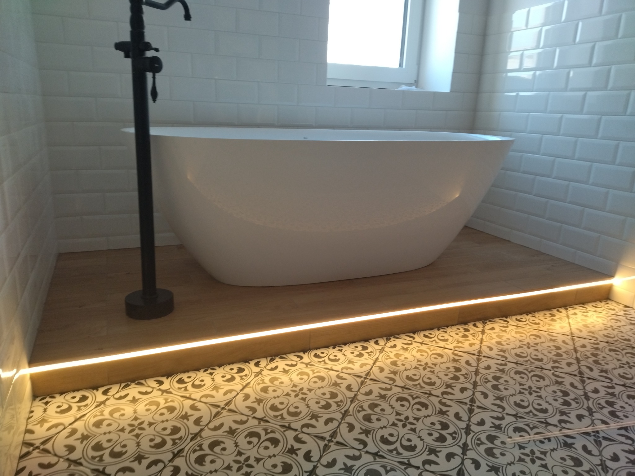 Nowoczesna łazienka z wolnostojącą wanną, białymi płytkami typu metro i podświetlaną podłogą z wzorzystymi płytkami. Czarna, wolnostojąca bateria wannowa.