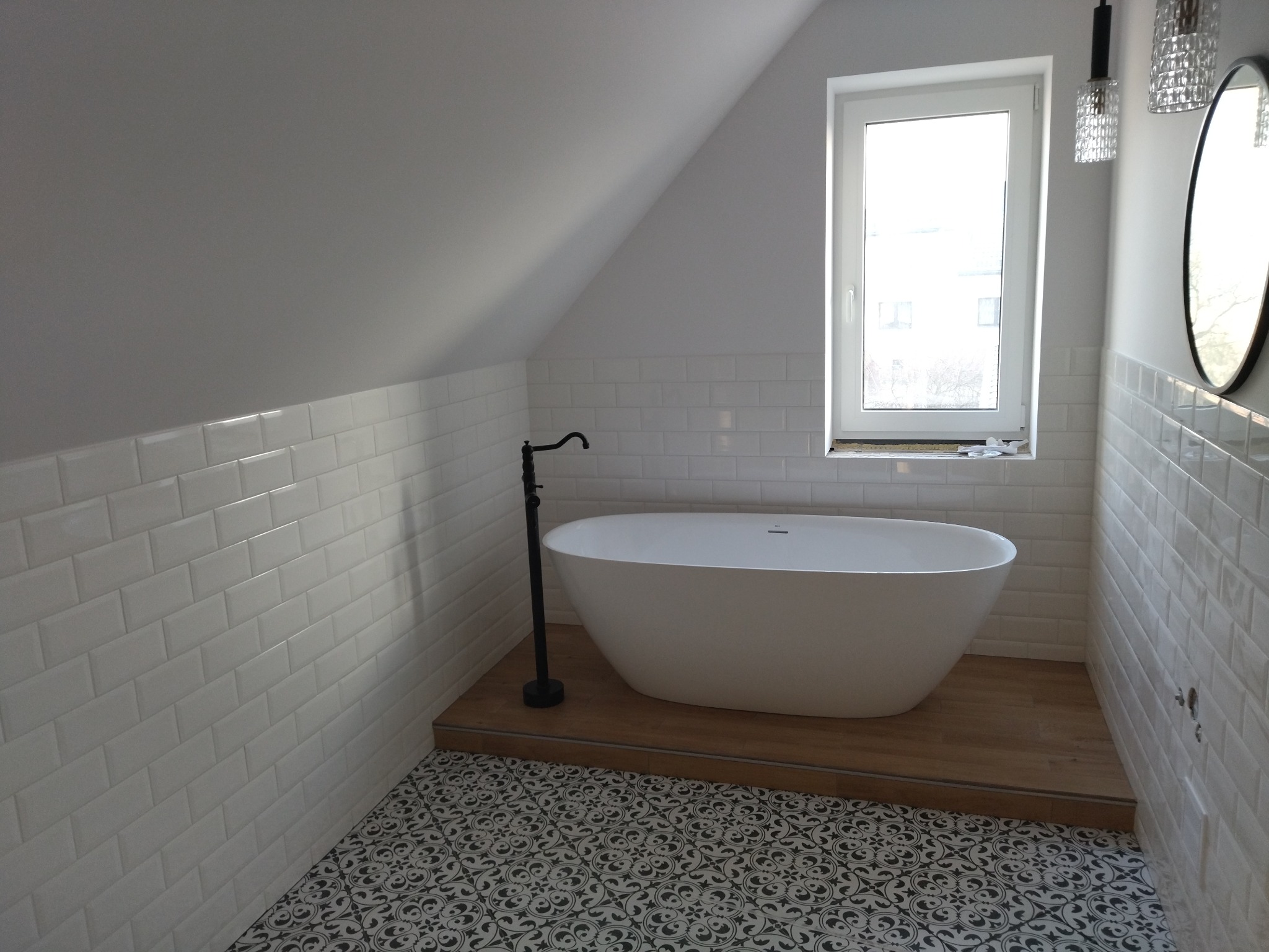 Biała wanna wolnostojąca na podeście z drewnopodobnych paneli, w łazience na poddaszu z białą cegiełką i wzorzystą podłogą. Nowoczesne oświetlenie i okrągłe lustro.