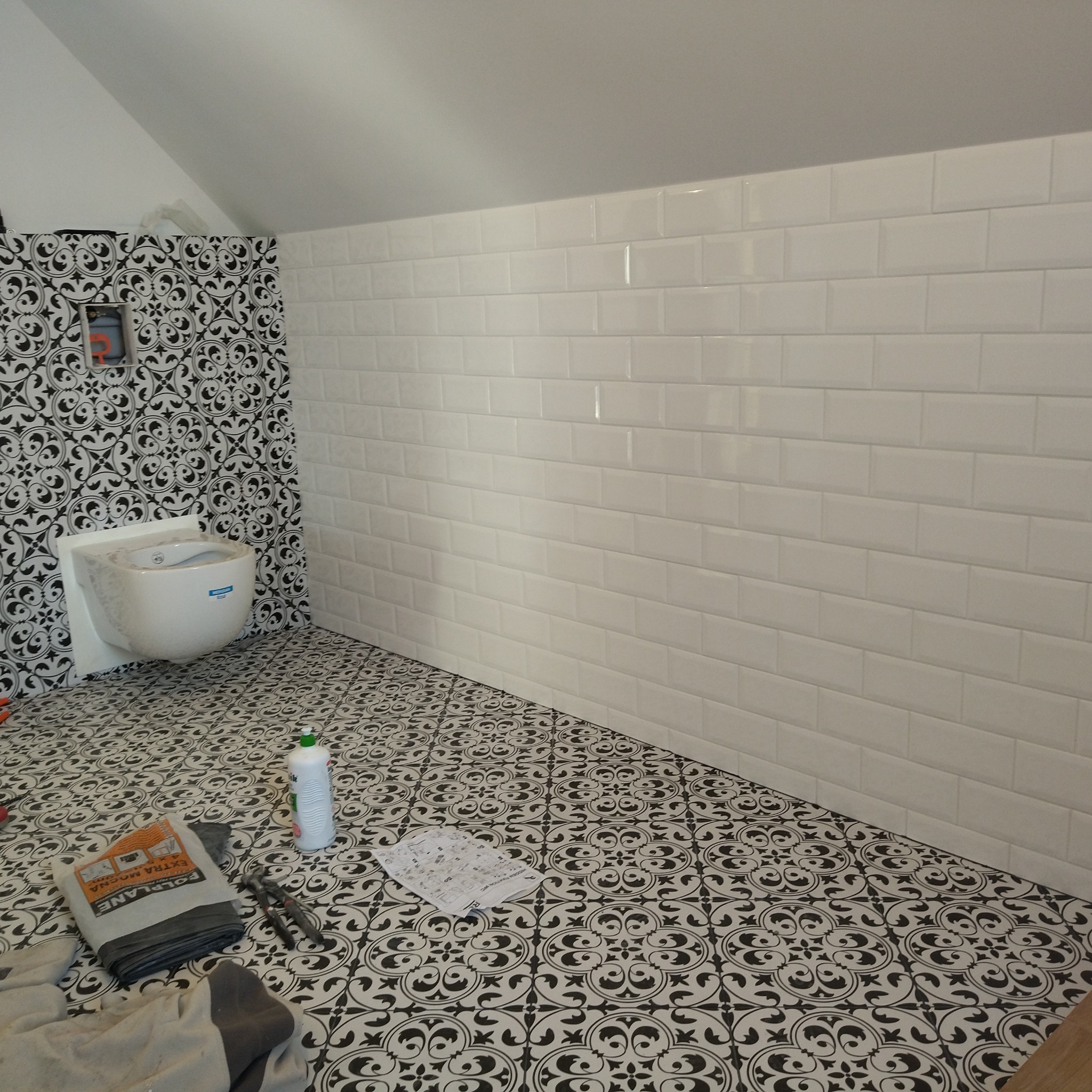 Wykończona łazienka na poddaszu z białą, cegiełkową glazurą i wzorzystymi płytkami podłogowymi. Umywalka zamontowana na ścianie z czarno-białą tapetą.