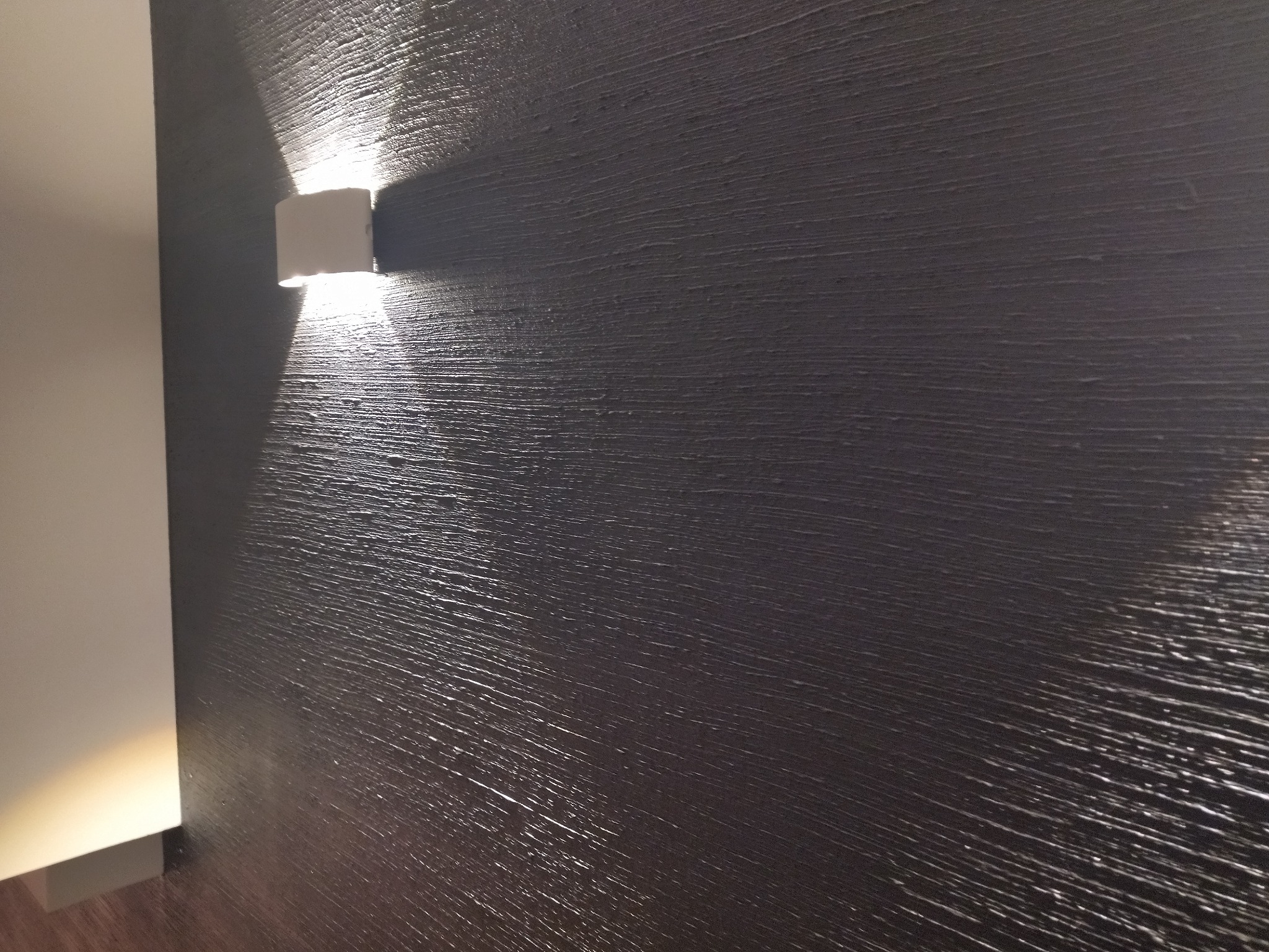 Szara ściana z wyraźną fakturą, oświetlona przez biały kinkiet, tworząca grę światła i cienia. Minimalistyczny design wnętrza.