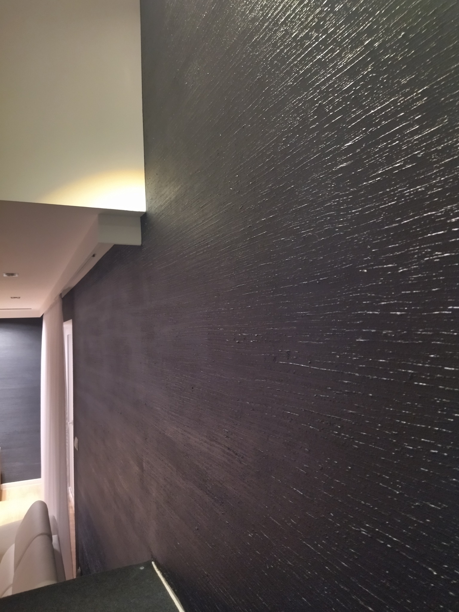 Czarna ściana z wyraźną fakturą, oświetlona od góry, widoczna z perspektywy. Fragment sufitu i wnętrza pokoju w tle. Dekoracyjne wykończenie ściany.