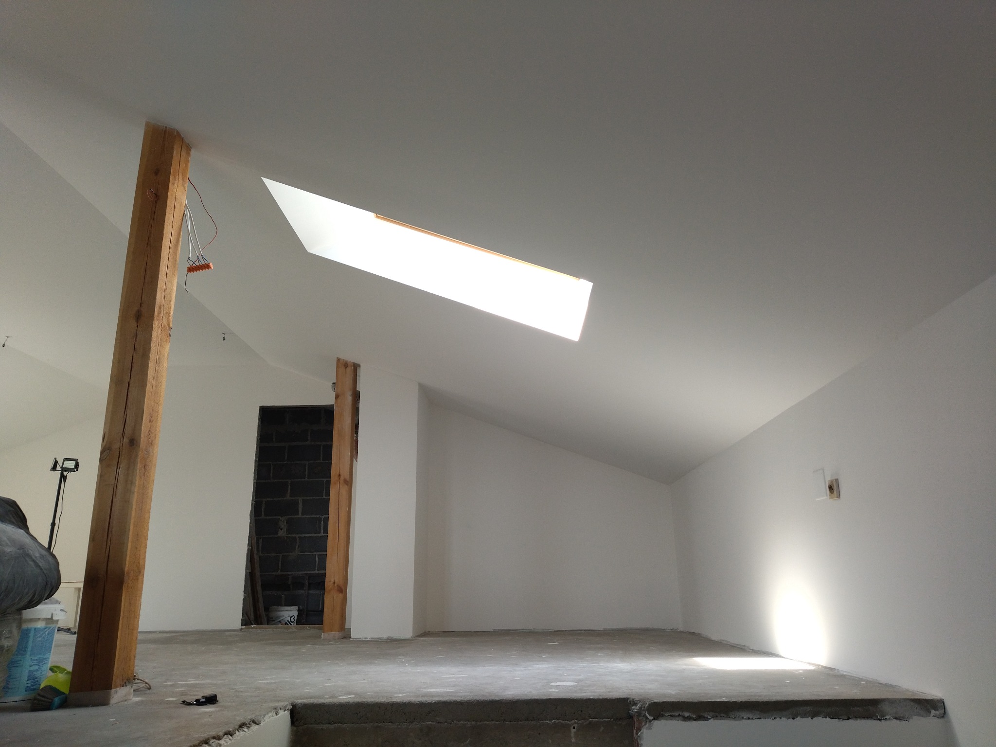 Wnętrze poddasza po malowaniu ścian na biało, z drewnianymi belkami i oknem dachowym. Widoczny surowy beton na podłodze i fragment instalacji elektrycznej.