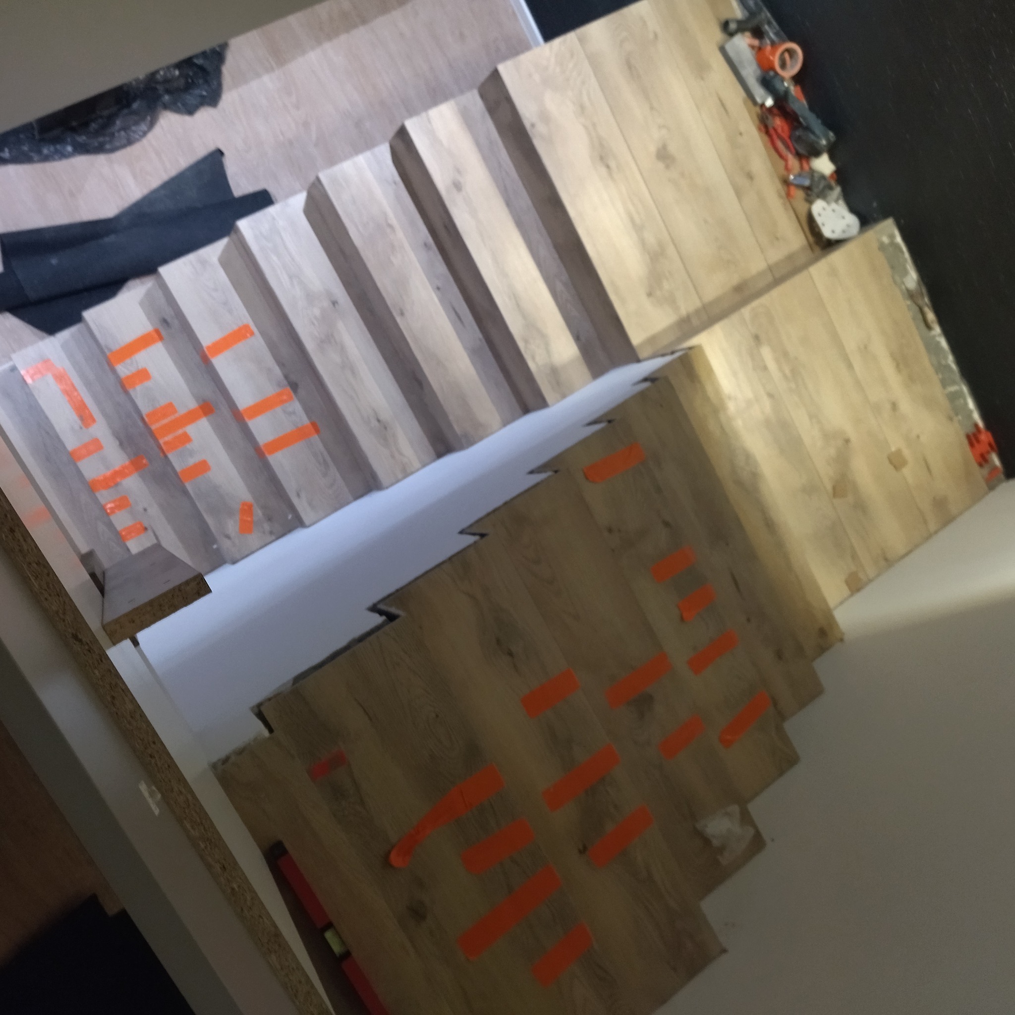 Montaż drewnopodobnych paneli na schodach z użyciem pomarańczowej taśmy. Widoczne narzędzia i materiały budowlane. Ujęcie z góry, w trakcie prac wykończeniowych.