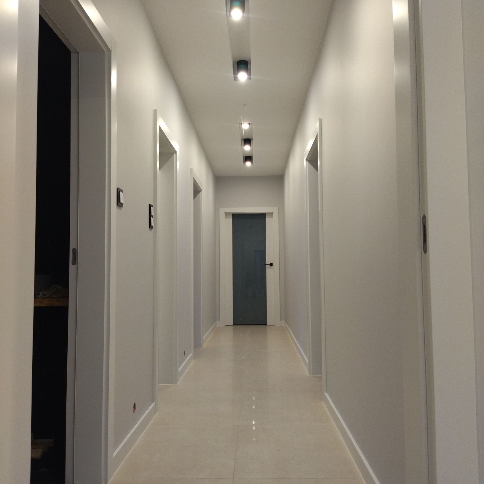 Długi, jasny korytarz z białymi drzwiami i punktowym oświetleniem sufitowym. Podłoga wyłożona jasnymi płytkami. Widok w głąb korytarza.