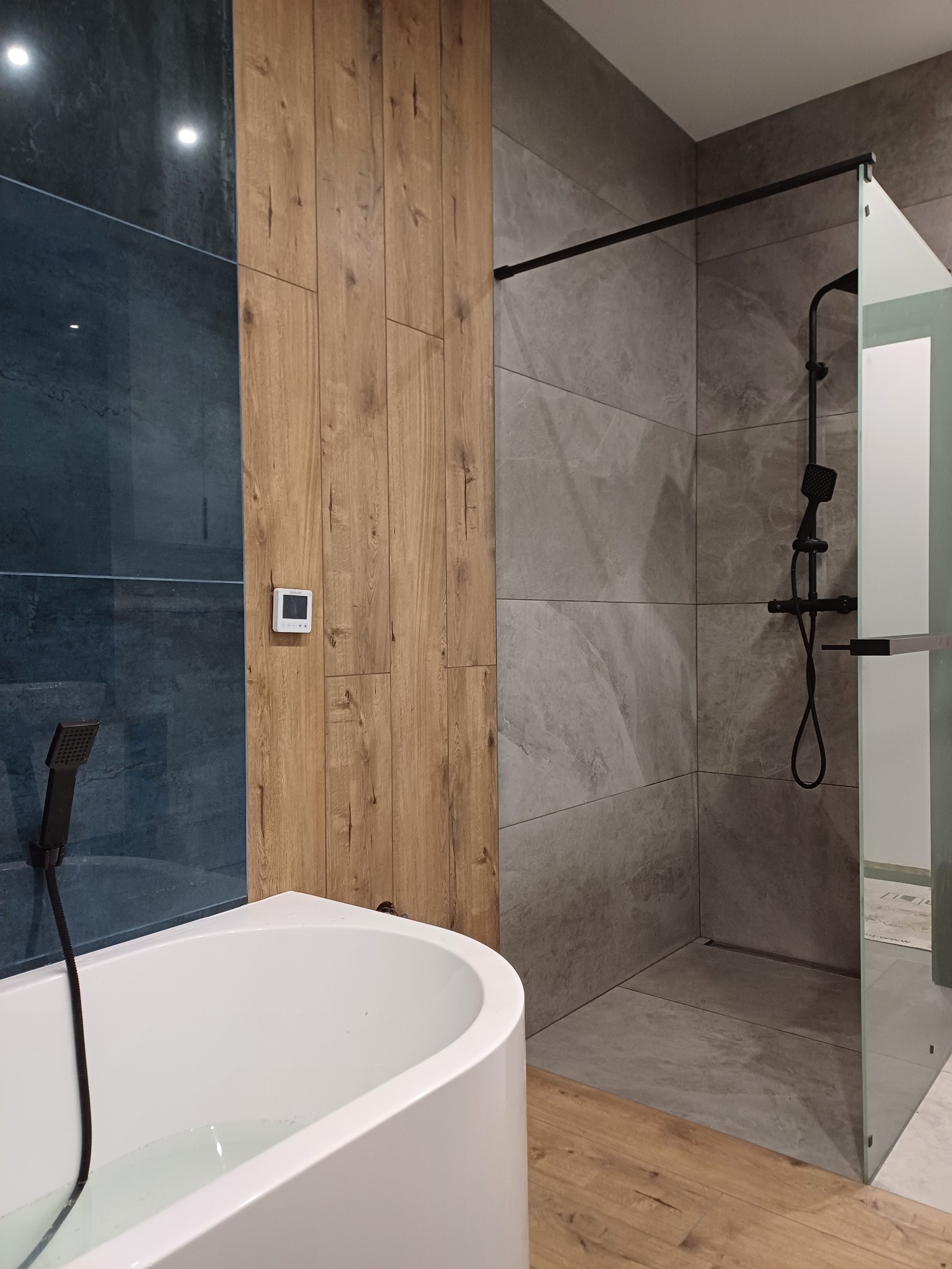 Nowoczesna łazienka z kabiną prysznicową typu walk-in, wanną i panelami ściennymi imitującymi drewno. Minimalistyczny design z czarnymi akcentami.