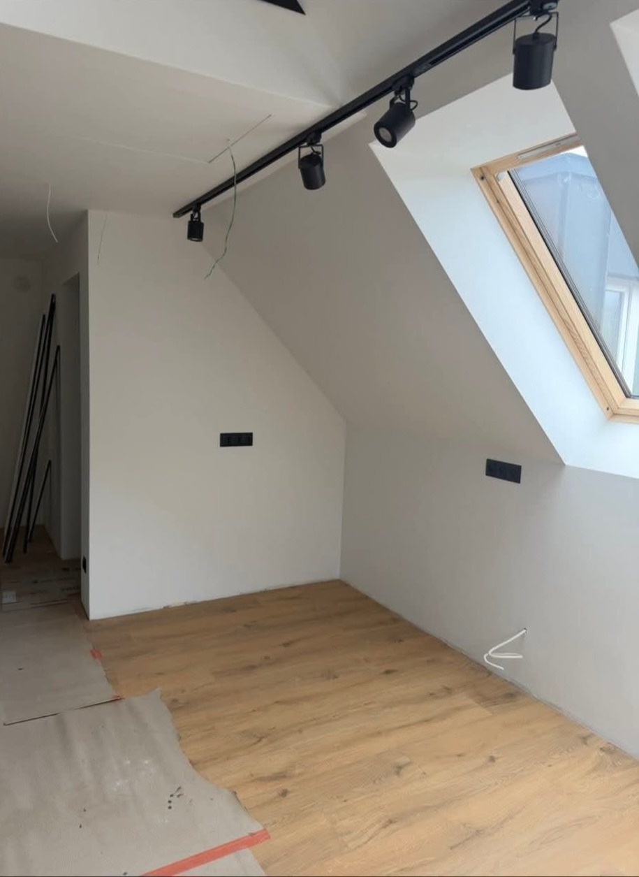 Pokój na poddaszu z oknem dachowym i drewnianą podłogą w trakcie układania. Widoczne listwy przypodłogowe i oświetlenie szynowe.