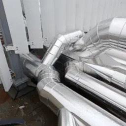 Metalowe rury izolacyjne, układane na zewnątrz budynku, widoczne łączenia i mocowania, wilgotna nawierzchnia, fragment konstrukcji wsporczej.