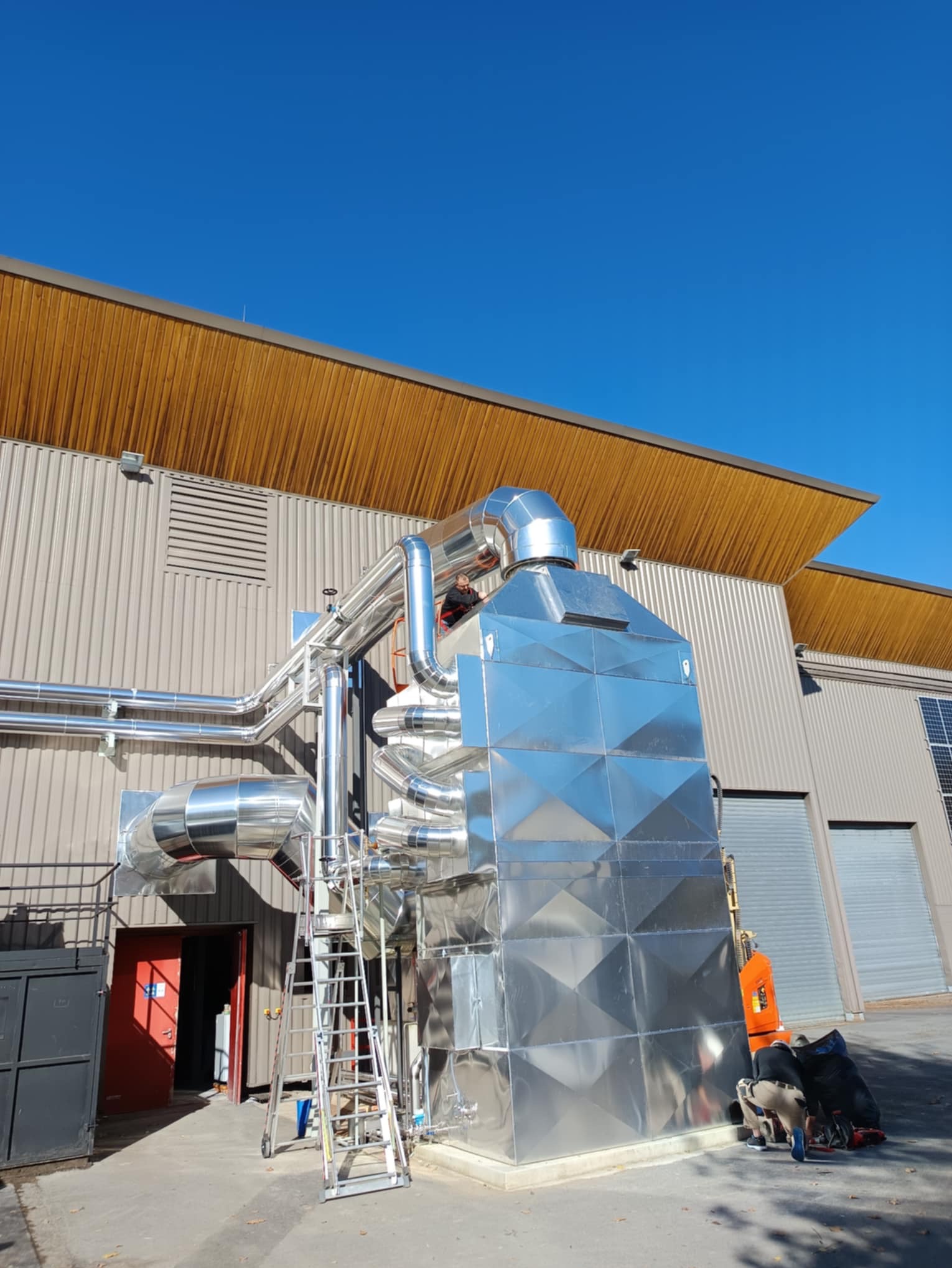 Ocieplenie przemysłowe budynku z blachy trapezowej. Widoczne rury i drabina, elementy systemu wentylacji. Osoba przy pracy. Jasne niebo i budynek z panelami słonecznymi w tle.