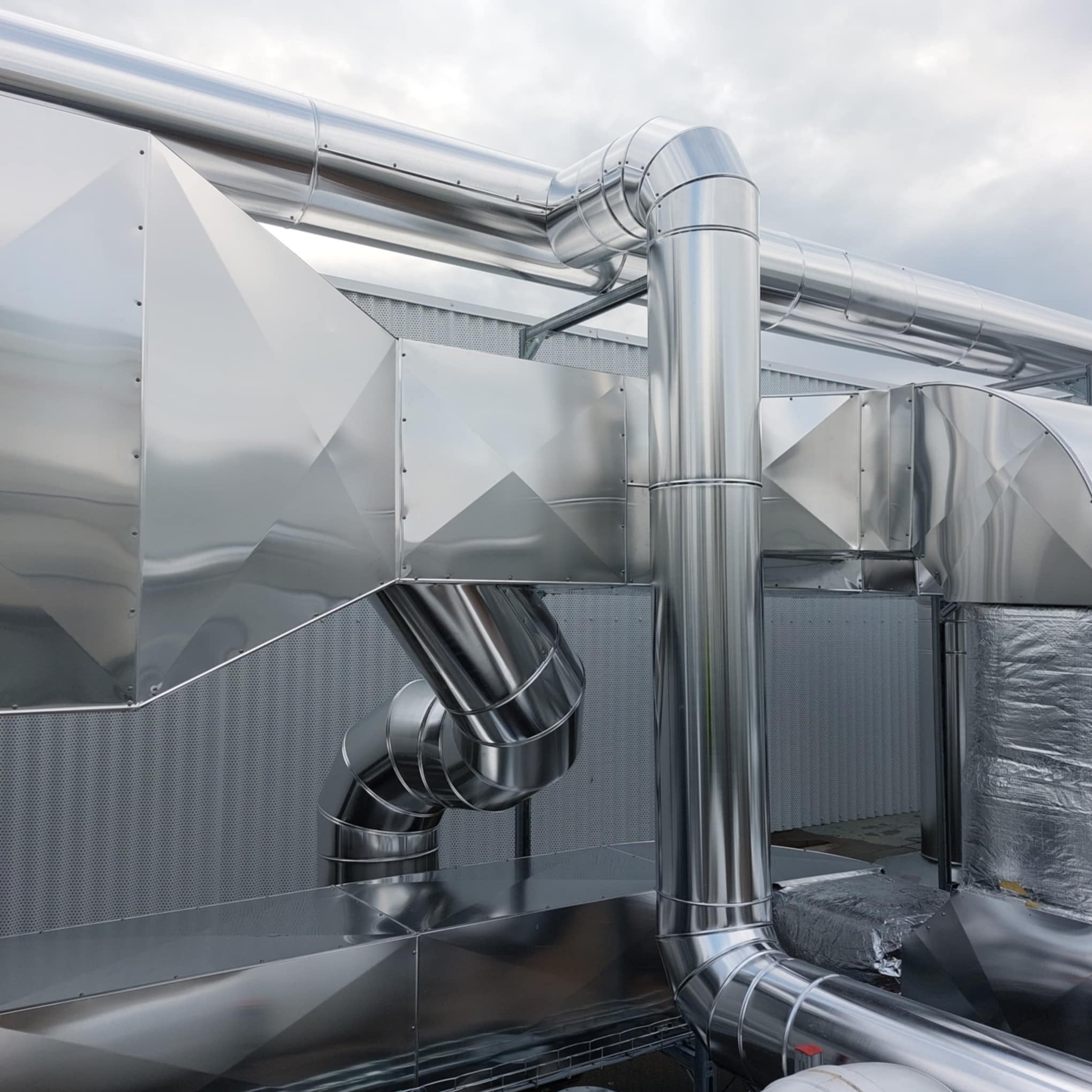 Błyszcząca instalacja wentylacyjna z metalu na tle szarego nieba. Widoczne rury, kolanka, elementy wentylacyjne i perforowana ściana. Detal techniczny.