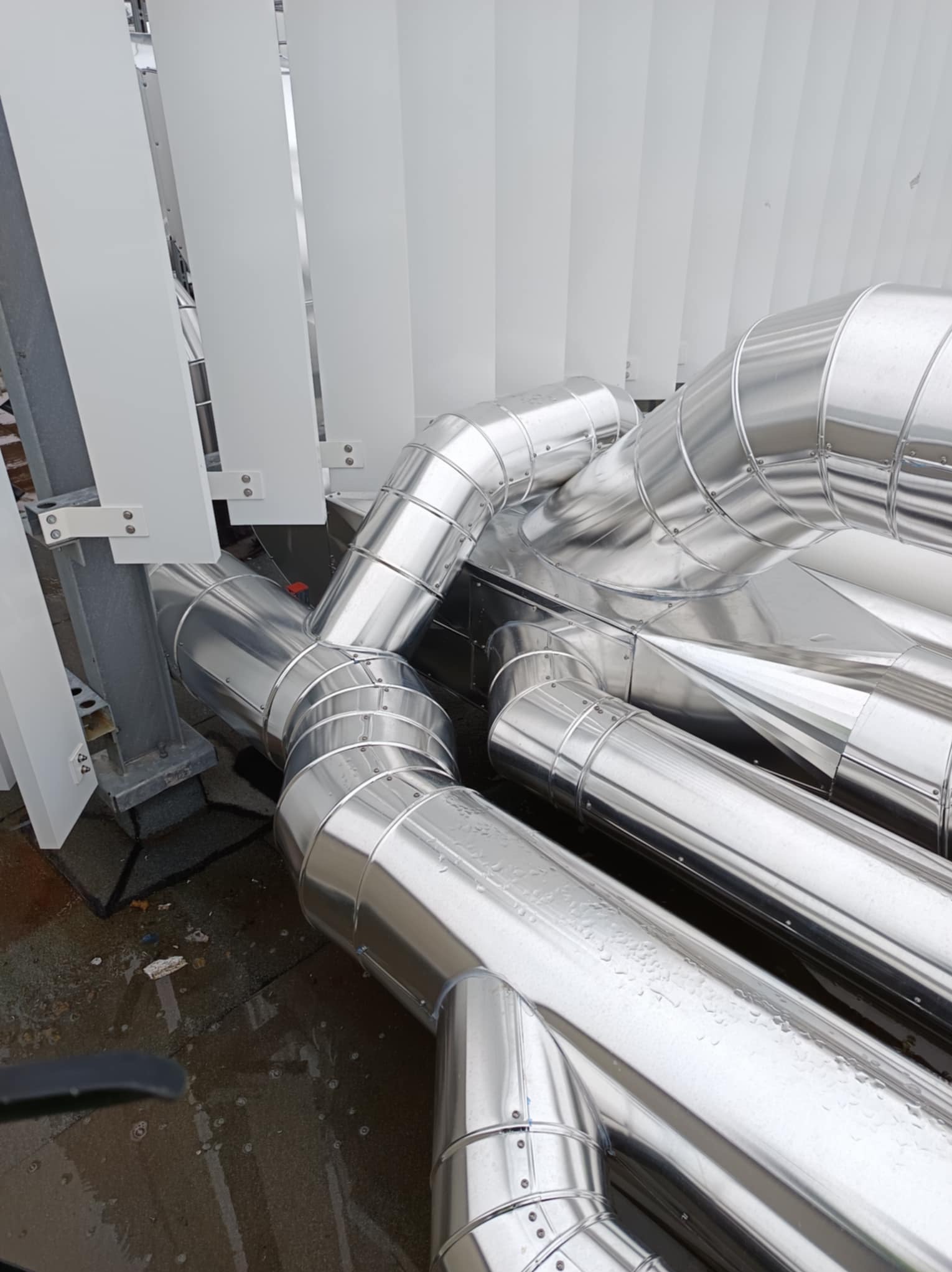 Metalowe rury izolacyjne, układane na zewnątrz budynku, widoczne łączenia i mocowania, wilgotna nawierzchnia, fragment konstrukcji wsporczej.