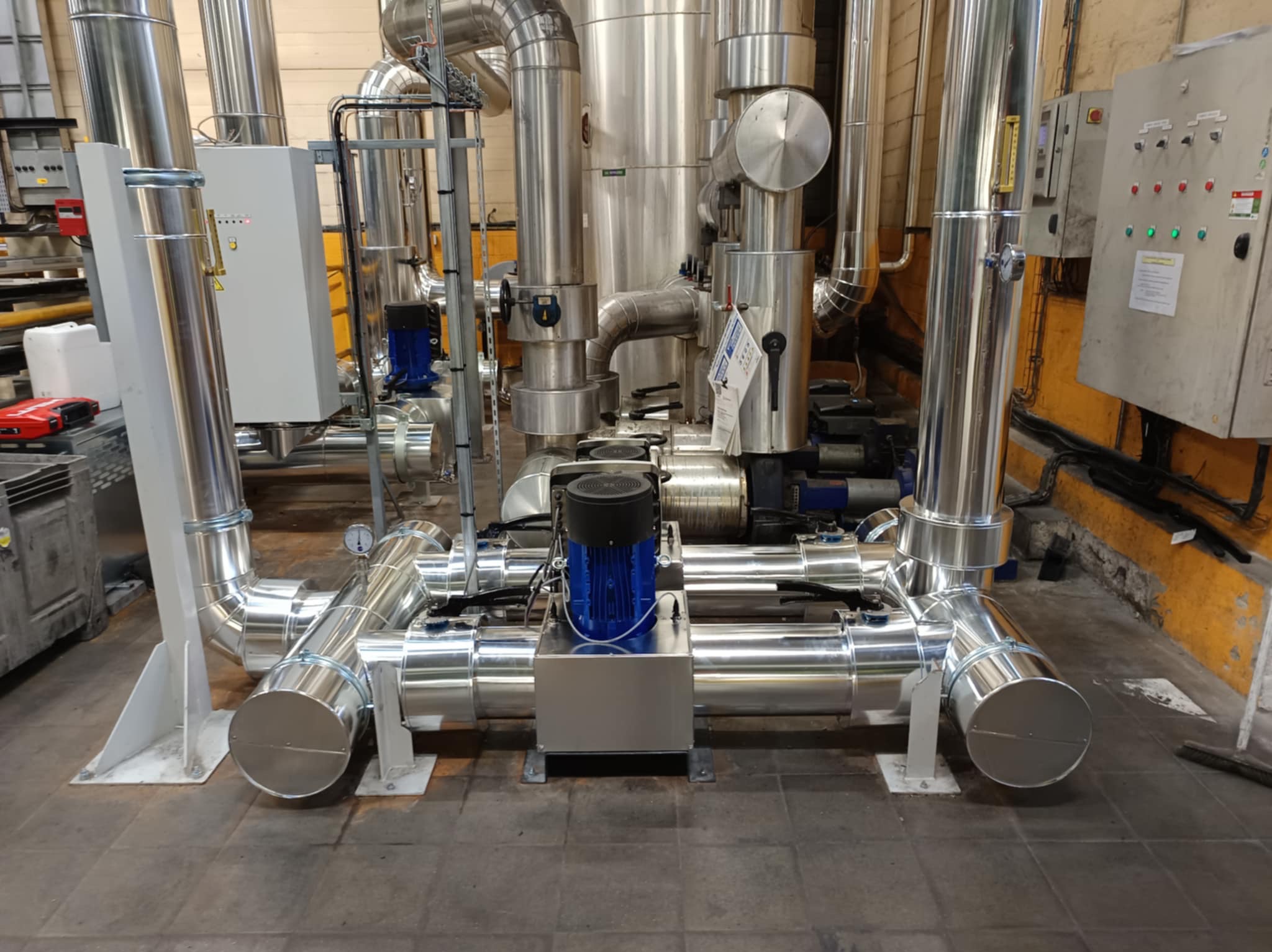 Izolacja przemysłowa: Błyszczące, metalowe rury i zbiorniki z widocznymi elementami montażowymi i niebieskimi pompami w hali przemysłowej.