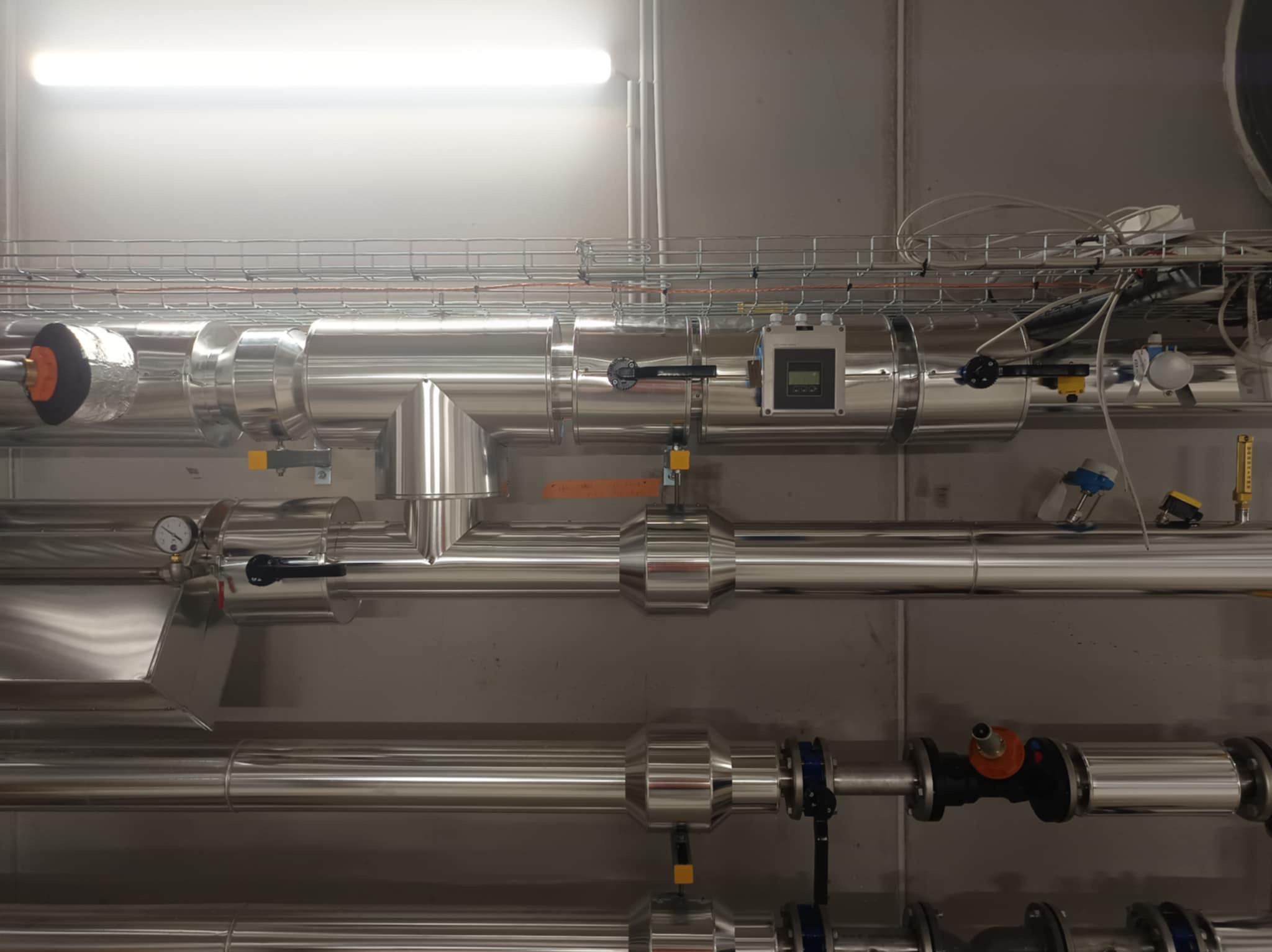 System rur z izolacją termiczną w pomieszczeniu technicznym, widoczne zawory, manometry i elementy sterujące. Metalowe rury z połyskiem.