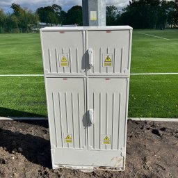 Amper-Spec - Nowa, dwuczęściowa szafka elektryczna na gruncie obok boiska sportowego. Na szafce widoczne naklejki ostrzegawcze o napięciu elektrycznym.