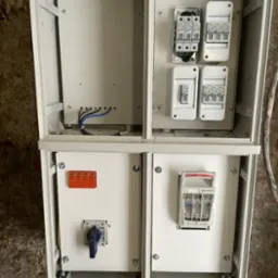 Otwarta, biała skrzynka elektryczna z bezpiecznikami, wyłącznikami i przewodami. Widoczne szczegóły montażu i elementy instalacji.