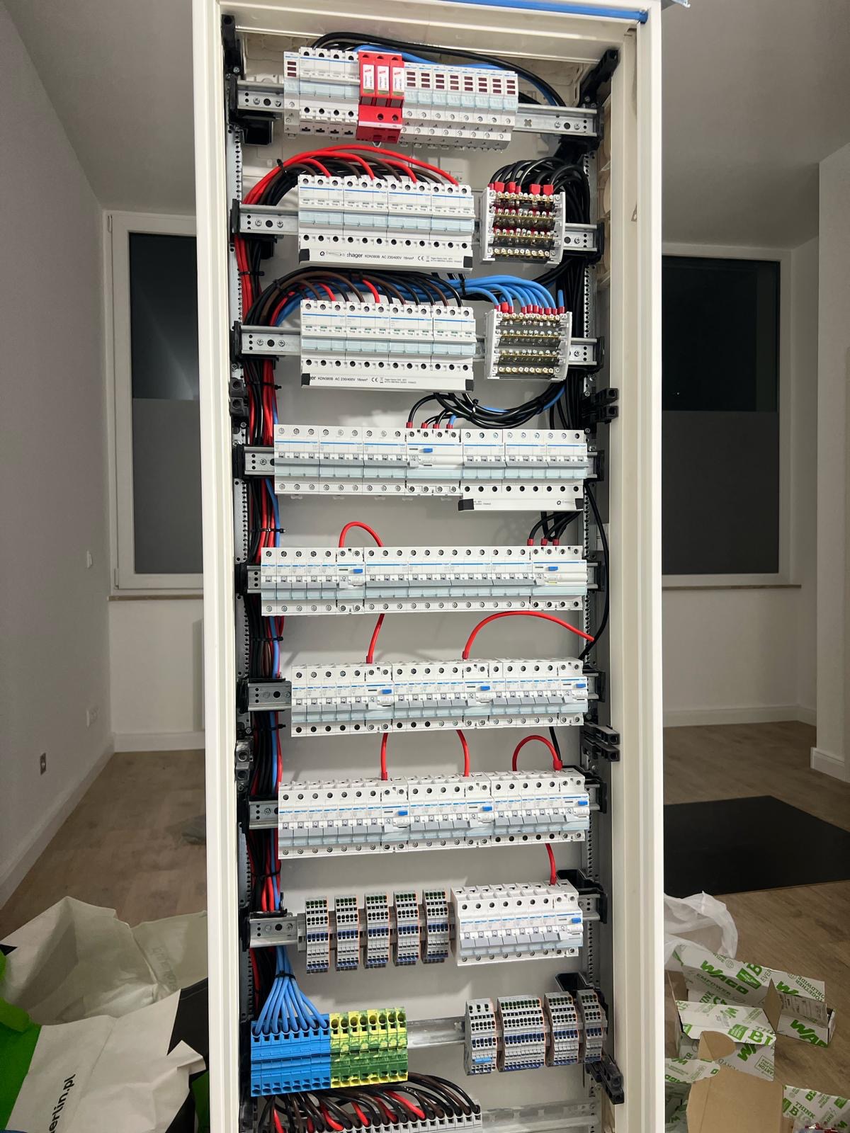 Rozdzielnica elektryczna wnękowa z licznymi zabezpieczeniami, przewodami i złączkami, zamontowana w jasnym pomieszczeniu z oknem, widok z bliska.