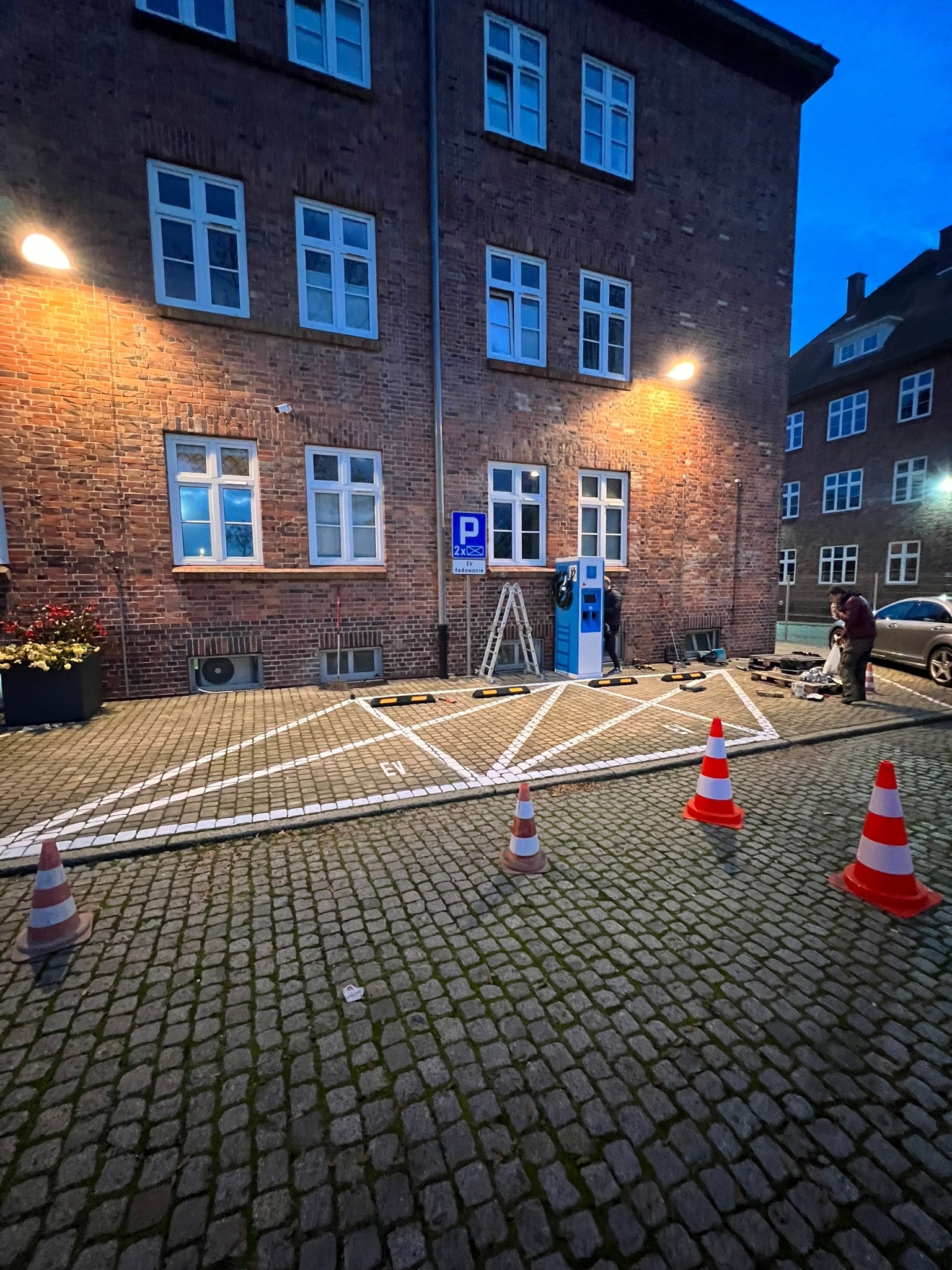 Montaż stacji ładowania pojazdów elektrycznych na parkingu z kostki brukowej, widoczni pracownicy i pomarańczowe pachołki.