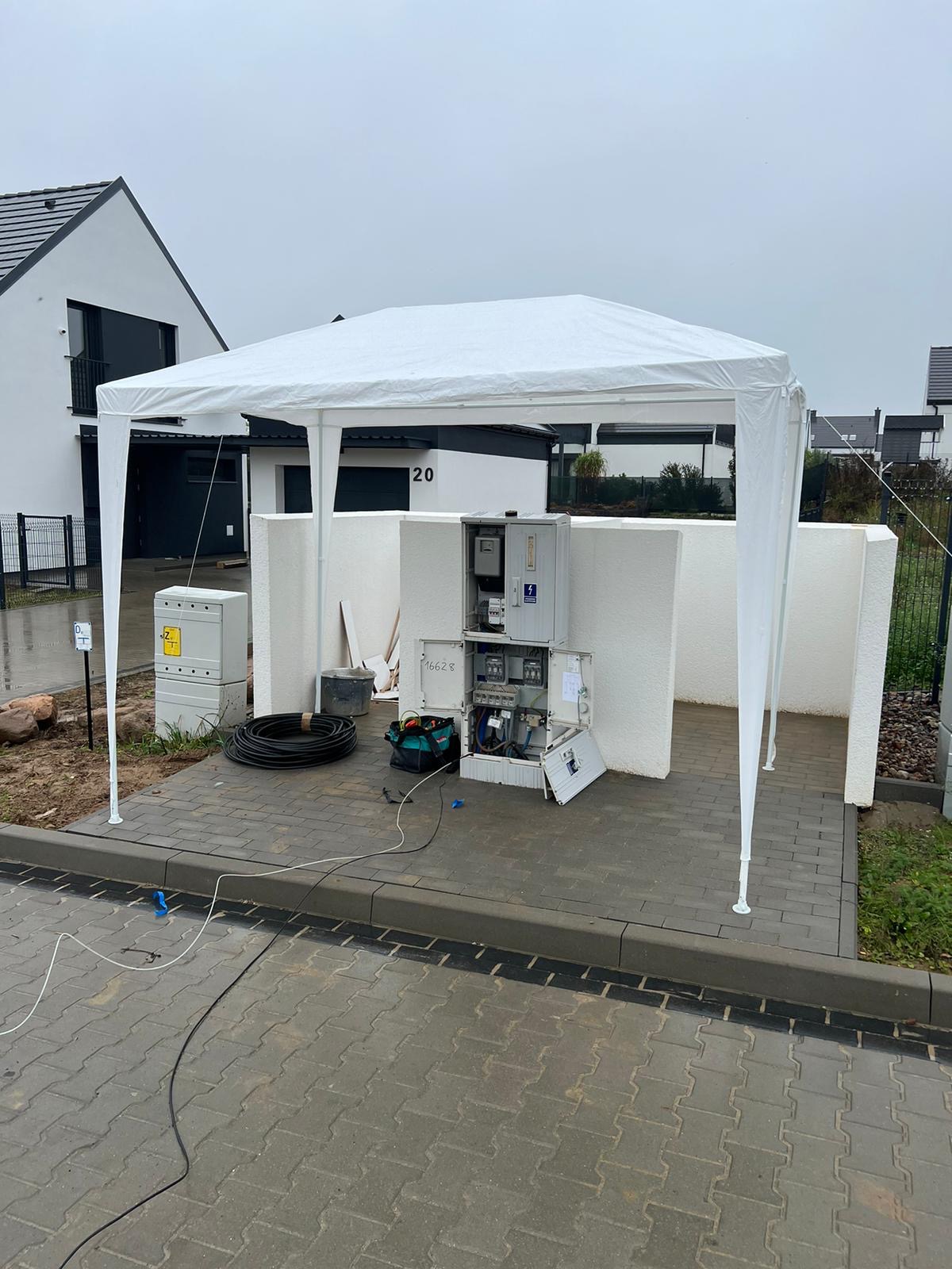 Otwarta skrzynka elektryczna z rozłożonymi bezpiecznikami i przewodami pod białym namiotem, na tle nowoczesnego domu jednorodzinnego.