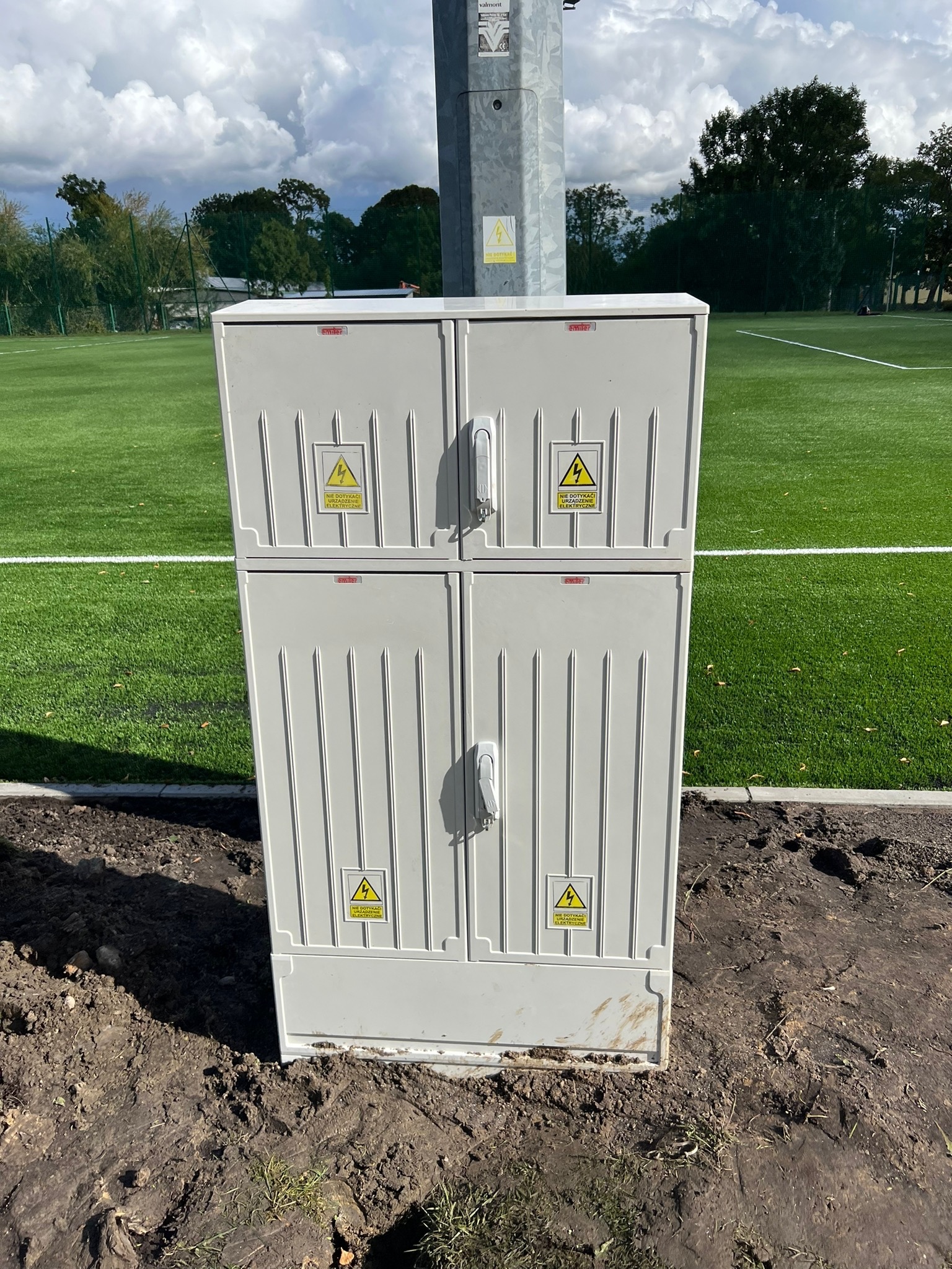 Nowa, dwuczęściowa szafka elektryczna na gruncie obok boiska sportowego. Na szafce widoczne naklejki ostrzegawcze o napięciu elektrycznym.