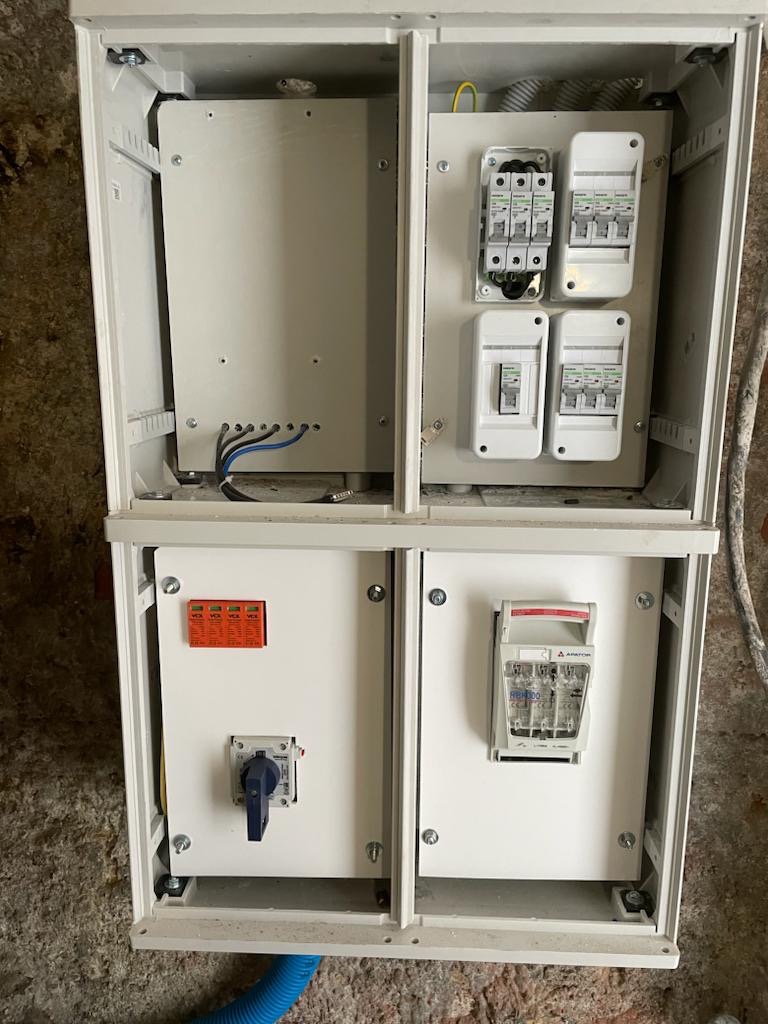 Otwarta, biała skrzynka elektryczna z bezpiecznikami, wyłącznikami i przewodami. Widoczne szczegóły montażu i elementy instalacji.