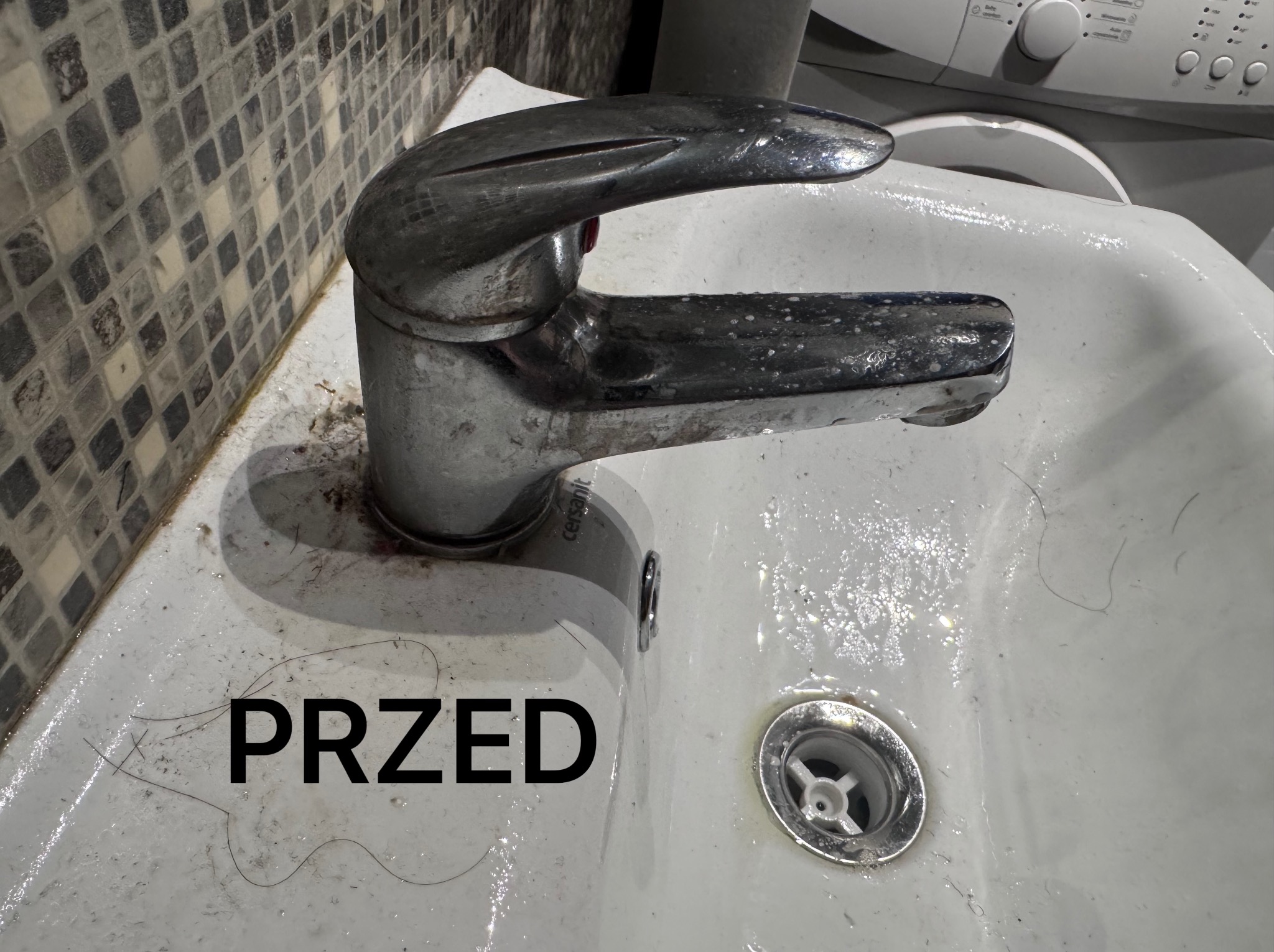 Zabrudzona umywalka z włosami i osadem, bateria pokryta plamami, widoczny napis 'PRZED', w tle pralka. Brak czystości i higieny.