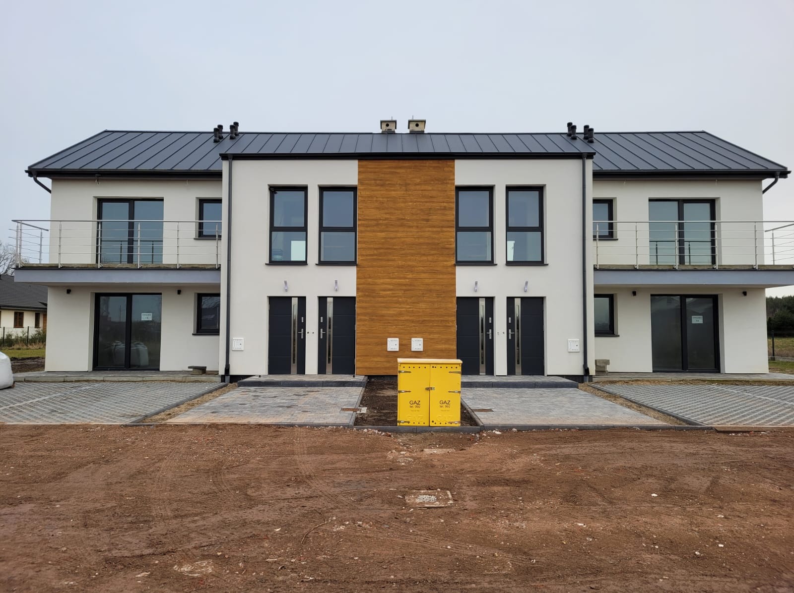 Nowoczesny bliźniak z białymi elewacjami i drewnianymi akcentami. Widoczne balkony z metalowymi balustradami i czarne drzwi. Skrzynka gazowa na pierwszym planie.