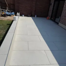 Jasne, prostokątne płyty tarasowe ułożone na zewnątrz budynku, z widocznymi elementami ogrodu i ceglanym murem w tle, oświetlone naturalnym światłem.