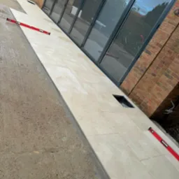 Układanie jasnych płytek na zewnątrz budynku, widoczne poziomice i odstępniki, częściowo ułożona powierzchnia, duże okna przesuwne w tle.