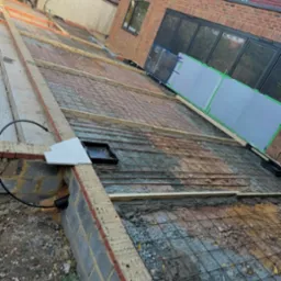 Zbrojenie pod wylewkę na tarasie z widoczną siatką, drewnianymi szalunkami i przygotowaną instalacją. Budynek w tle z oknami i drzwiami balkonowymi.