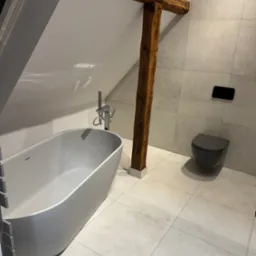 Nowoczesna łazienka na poddaszu z wanną wolnostojącą, szarą armaturą i drewnianymi belkami stropowymi. Minimalistyczny design, jasne płytki podłogowe i ścienne.