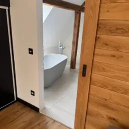 Nowoczesna łazienka z wolnostojącą wanną, drewnianymi belkami stropowymi i przesuwnymi drzwiami z drewna, widoczna z perspektywy pokoju z drewnianą podłogą.