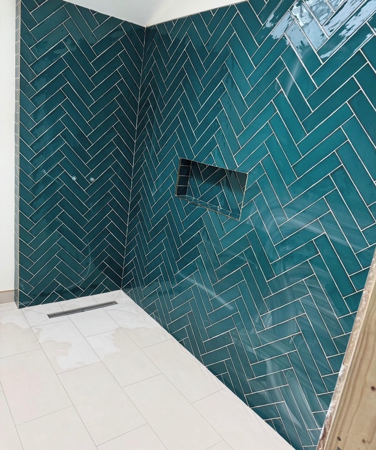 Wykończona łazienka z turkusową glazurą ułożoną w jodełkę i wnęką. Białe płytki podłogowe z odpływem liniowym. Widoczny fragment drewnianej konstrukcji.