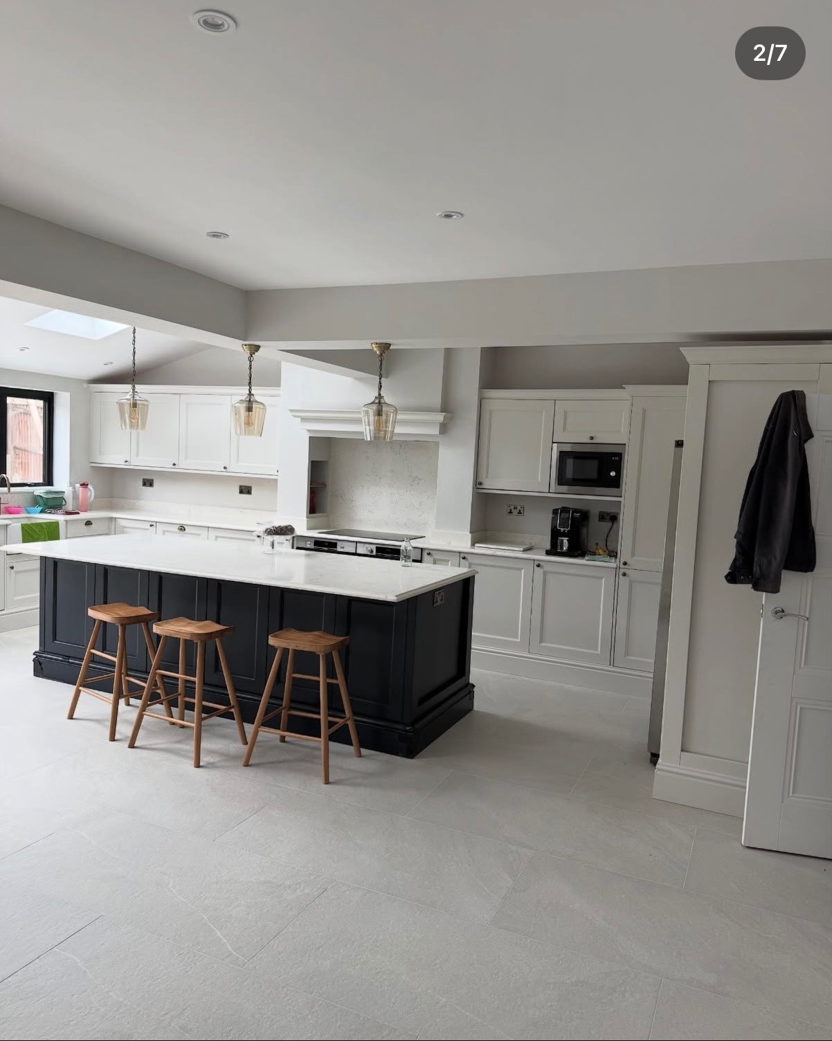 Jasna kuchnia po remoncie: białe szafki, czarna wyspa z marmurowym blatem, trzy drewniane stołki barowe, płytki podłogowe w odcieniach szarości.