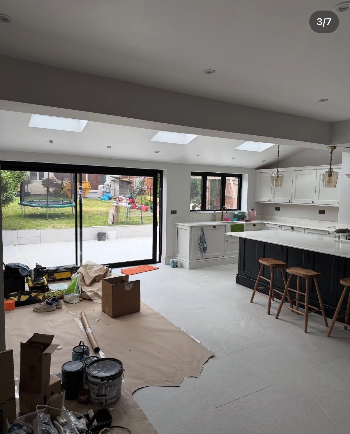 Wnętrze po remoncie: jasna kuchnia z wyspą i wyjściem na ogród. Widoczne narzędzia i farby wskazują na prace wykończeniowe.