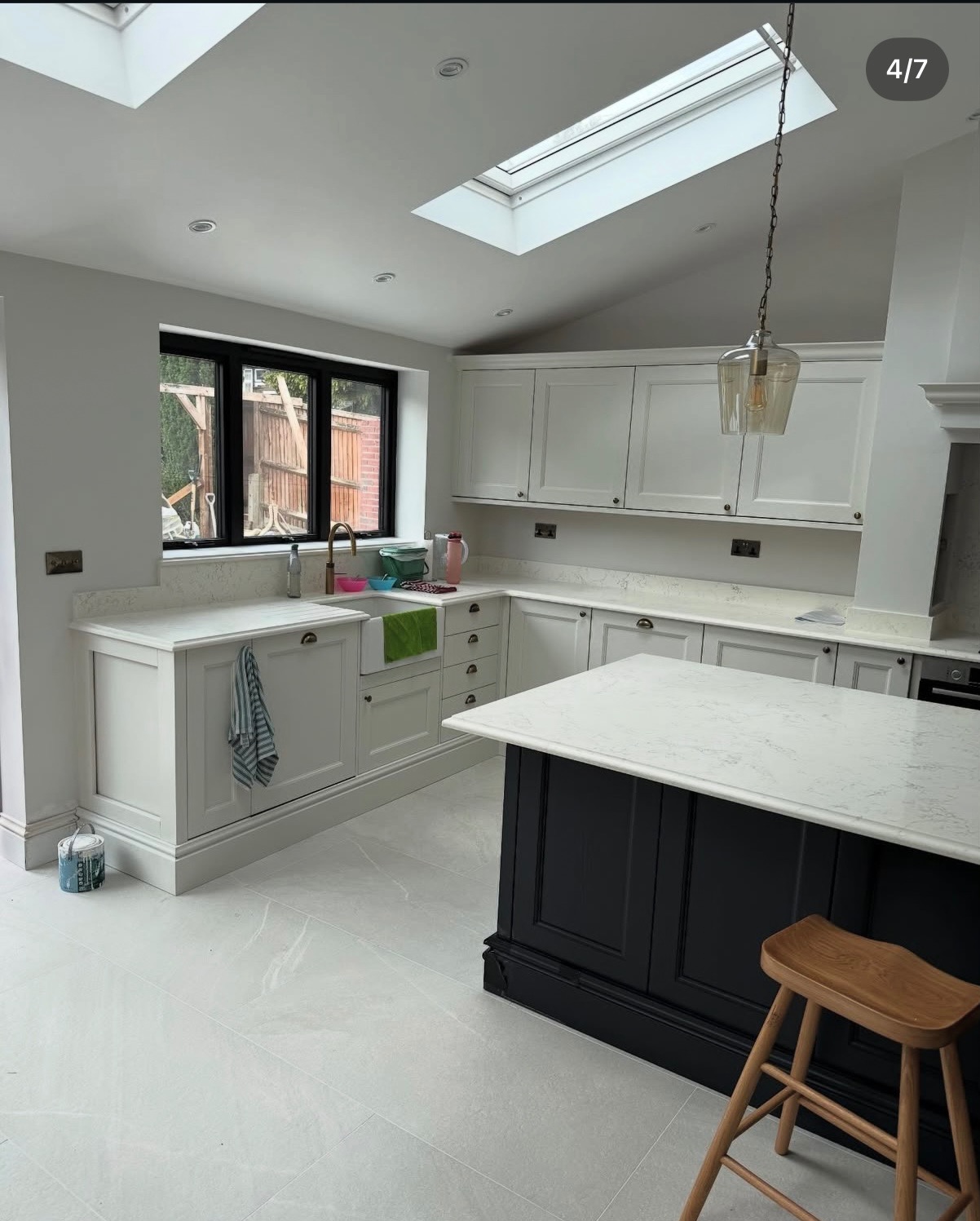Jasna kuchnia po remoncie: białe szafki, czarna wyspa z marmurowym blatem, okno i świetlik dachowy. Minimalistyczny styl, drewniany stołek.
