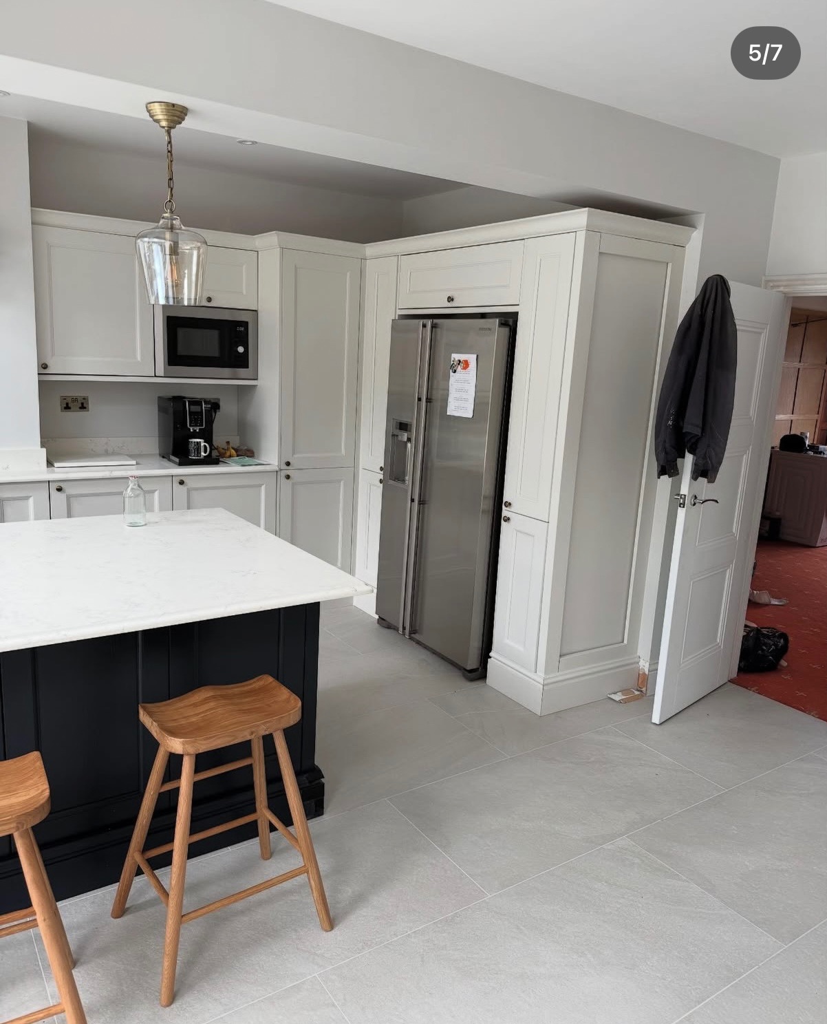 Jasna kuchnia z wyspą, drewnianymi stołkami i szarą podłogą z płytek. Widoczna zabudowa kuchenna, lodówka i lampa wisząca. Minimalistyczny design.