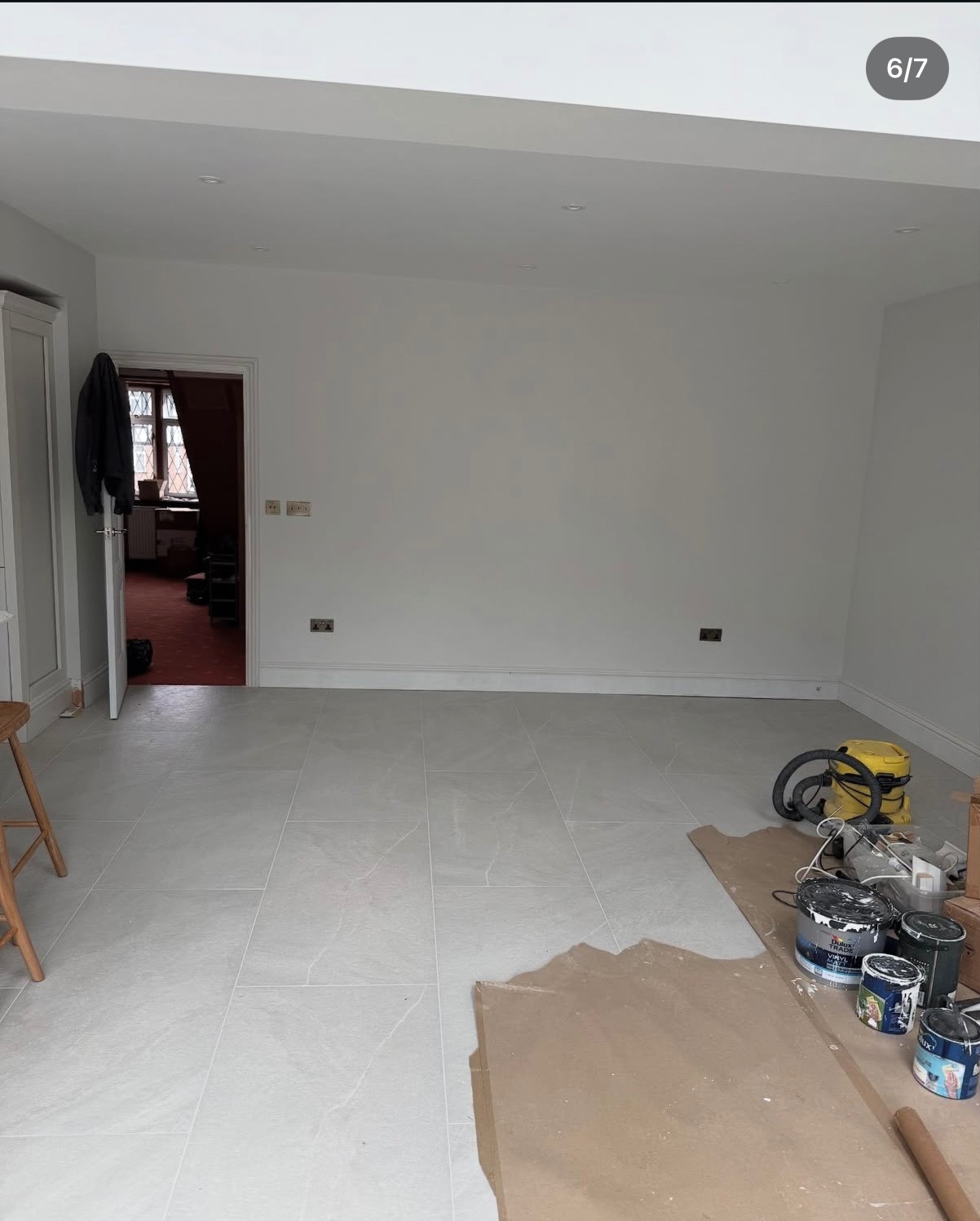 Jasne płytki podłogowe w odnowionym wnętrzu, z widocznymi puszkami farb i odkurzaczem, sugerującymi prace wykończeniowe. Minimalistyczny wystrój i świeże wykończenie.