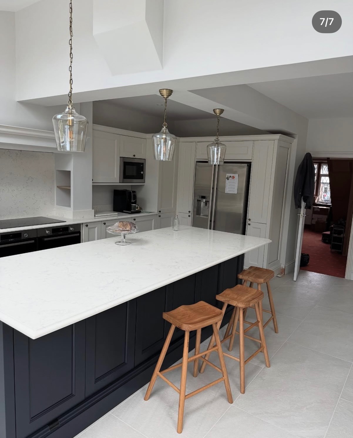 Elegancka, jasna kuchnia z wyspą i drewnianymi stołkami barowymi. Wiszące lampy szklane dodają uroku. Minimalistyczny design z elementami klasycznymi.