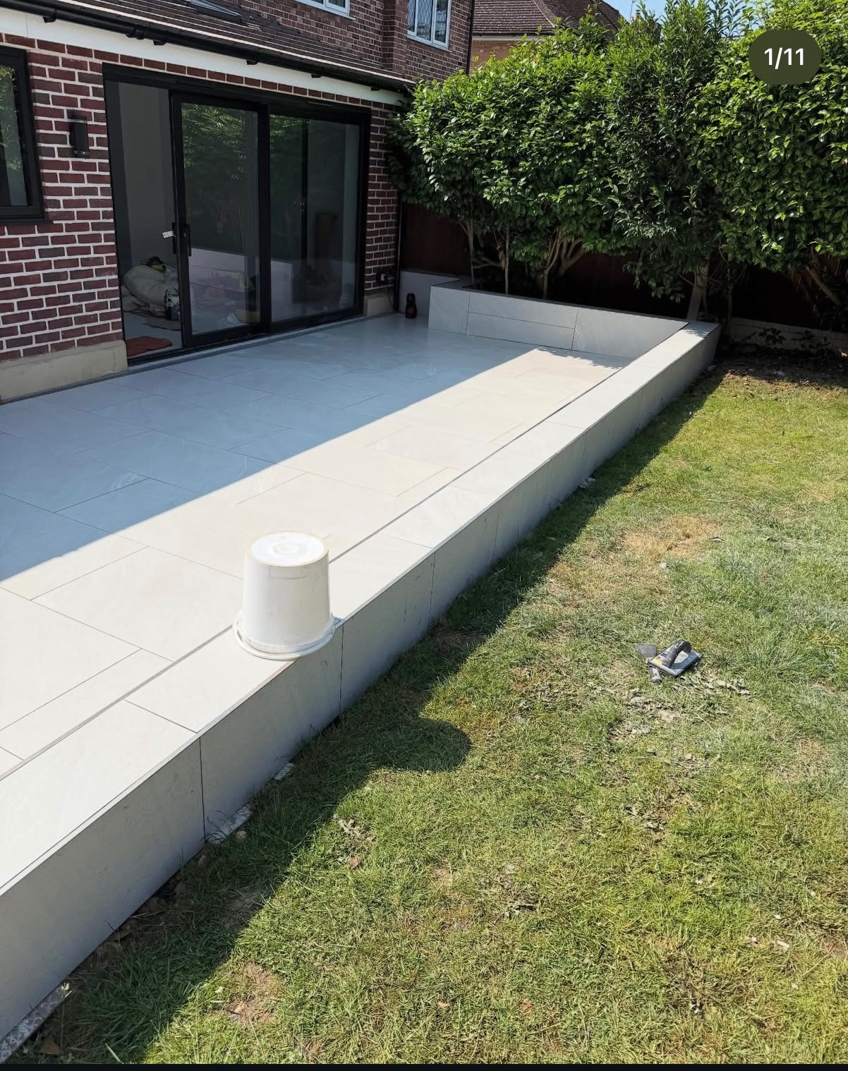 Jasne płytki tarasowe ułożone przy domu z cegły, z widokiem na zadbany trawnik i krzewy. Białe wiaderko na krawędzi tarasu, narzędzia na trawie.