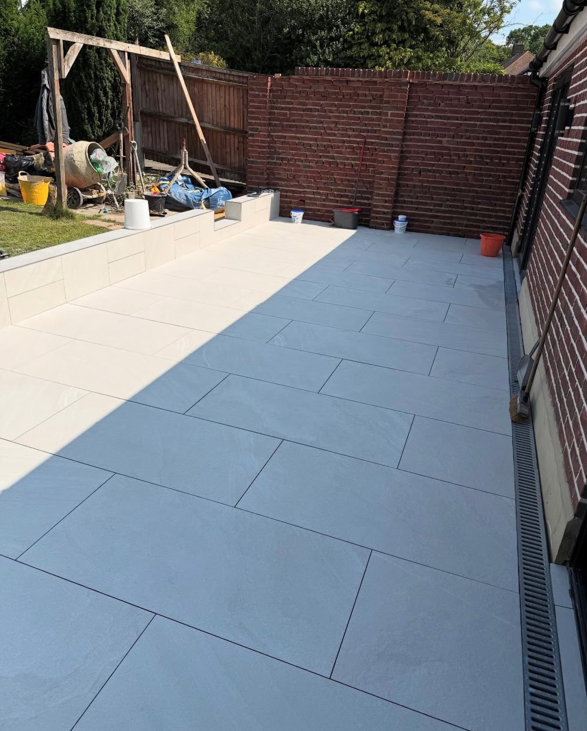 Wyłożony jasnoszarymi płytkami taras z odpływem liniowym przy ścianie budynku. W tle mur z cegły i drewniany płot. Widoczny sprzęt budowlany.