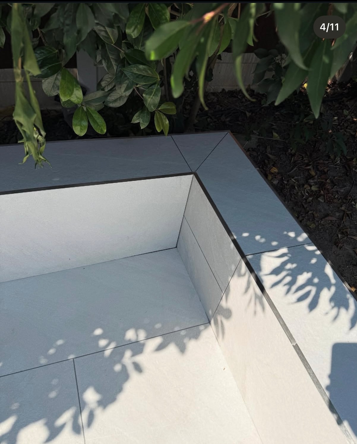 Wykończona, jasnoszara, kaskadowa konstrukcja z płytek ceramicznych w ogrodzie, z widocznymi cieniami liści i roślinnością w tle. Estetyczne ułożenie płytek.