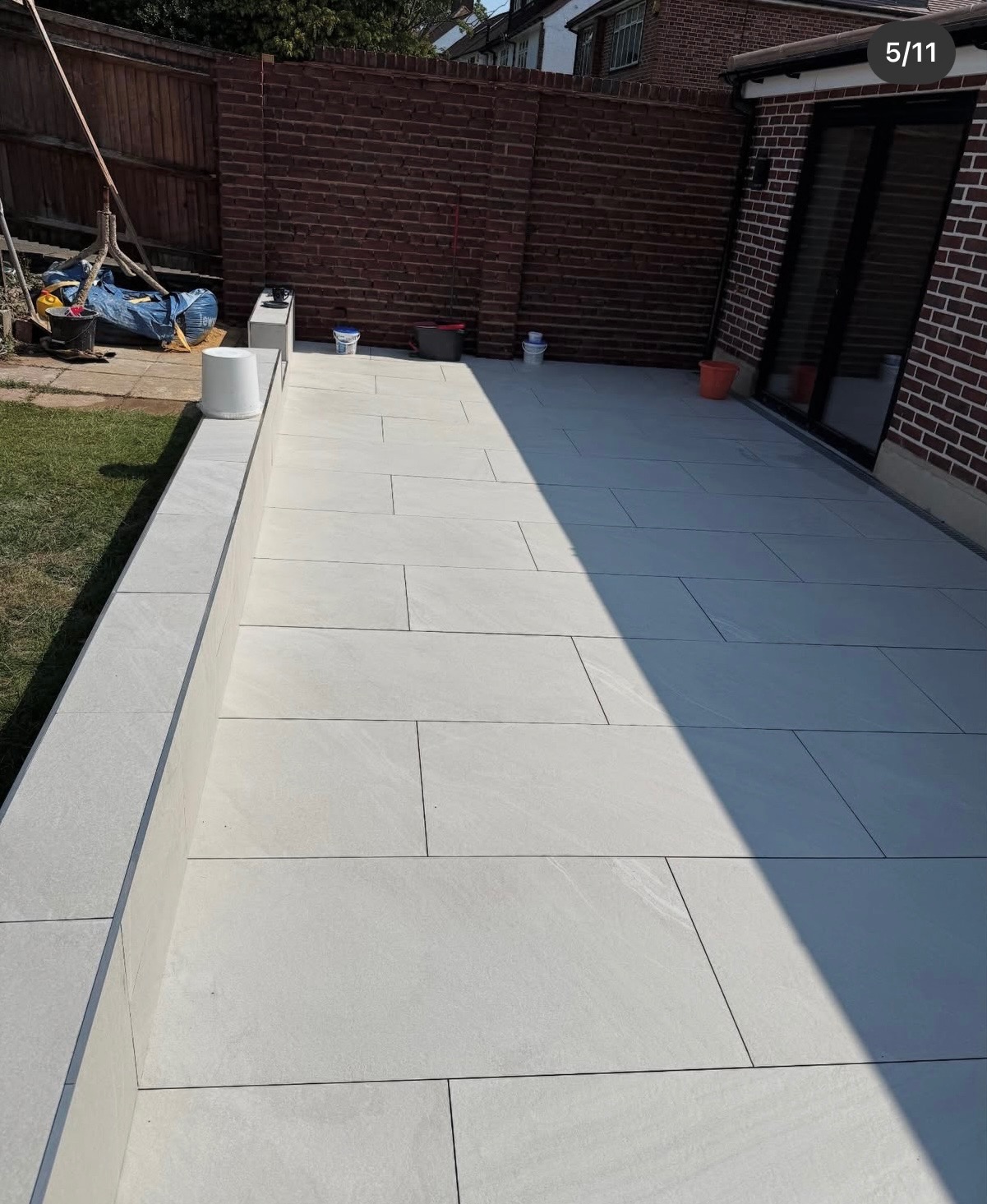 Jasne, prostokątne płyty tarasowe ułożone na zewnątrz budynku, z widocznymi elementami ogrodu i ceglanym murem w tle, oświetlone naturalnym światłem.