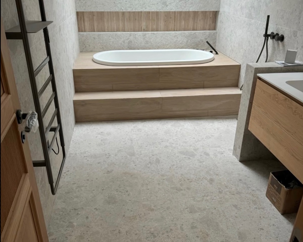 Elegancka łazienka z wanną na podwyższeniu wykończonym drewnem, szare płytki na podłodze i ścianach, oraz drabinka na ręczniki przy drewnianych drzwiach.