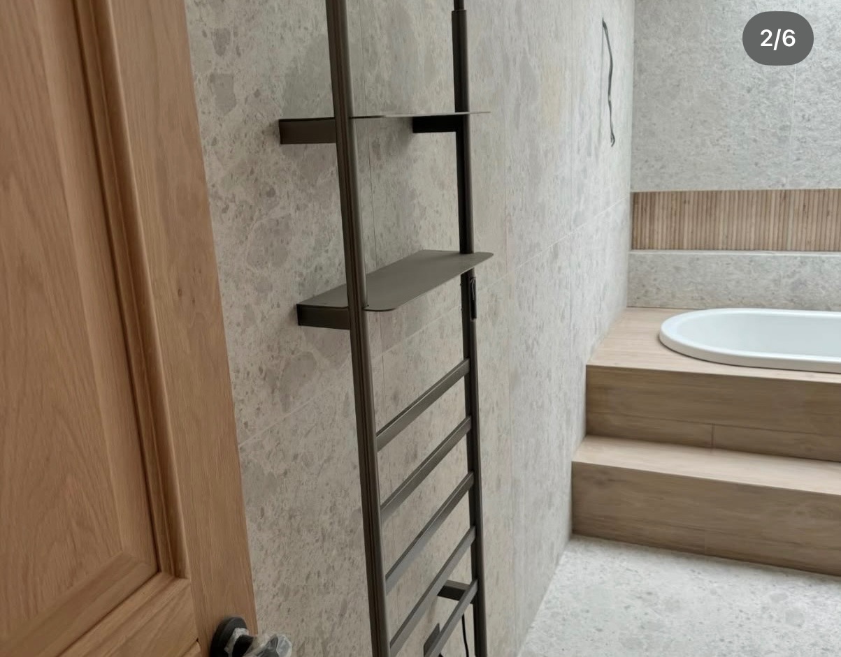 Wykończona łazienka z wanną na podeście, szarą płytką na ścianach i drewnianymi akcentami. Widoczny metalowy regał z półkami i fragment drewnianych drzwi.