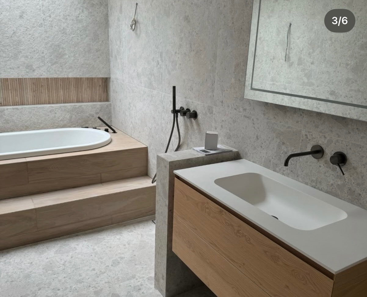 Nowoczesna łazienka z wanną na podeście wyłożonym drewnopodobnymi płytkami, umywalką z szafką i szarą, marmurkową okładziną ścienną.