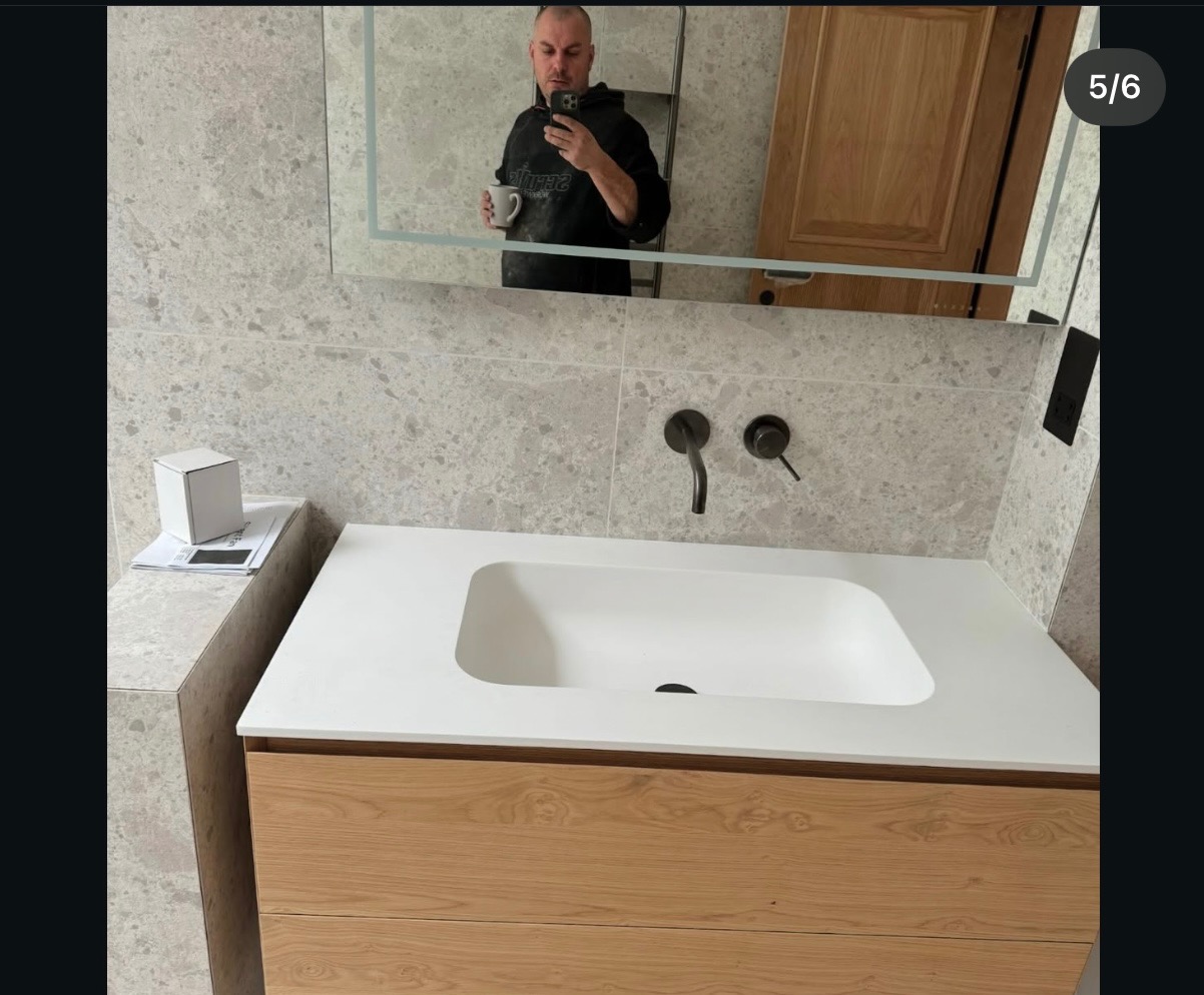 Nowoczesna łazienka z umywalką nablatową na drewnianej szafce, szarą glazurą i czarną armaturą. W lustrze odbija się osoba robiąca zdjęcie.