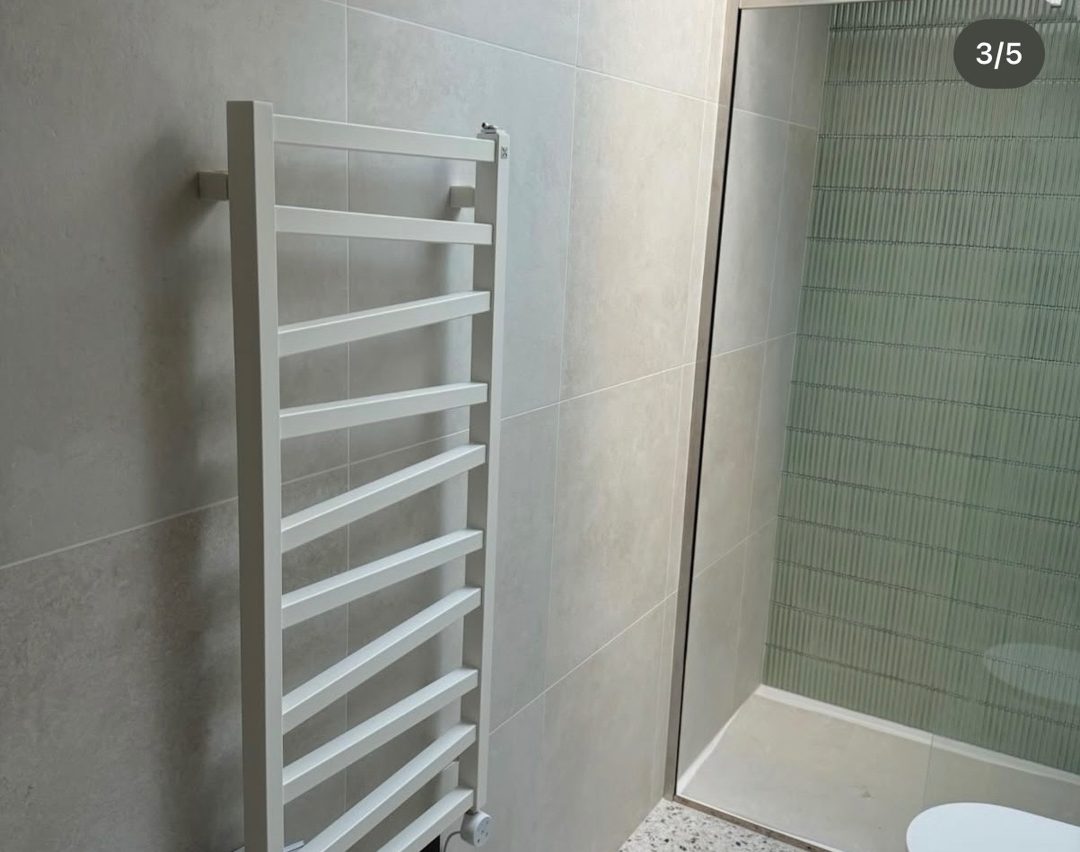 Biały grzejnik drabinkowy na tle szarej, płytkowej ściany w łazience z kabiną prysznicową z zielonymi, pionowymi płytkami. Nowoczesny design.