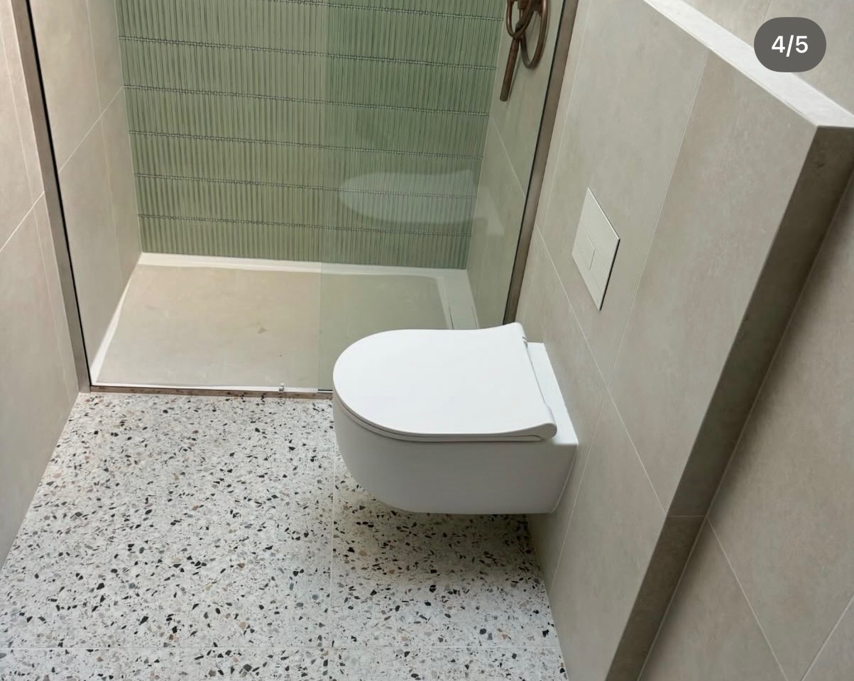 Nowoczesna łazienka po remoncie: podwieszana toaleta, szklana kabina prysznicowa z zielonymi płytkami, podłoga z lastryko i beżowe płytki ścienne.