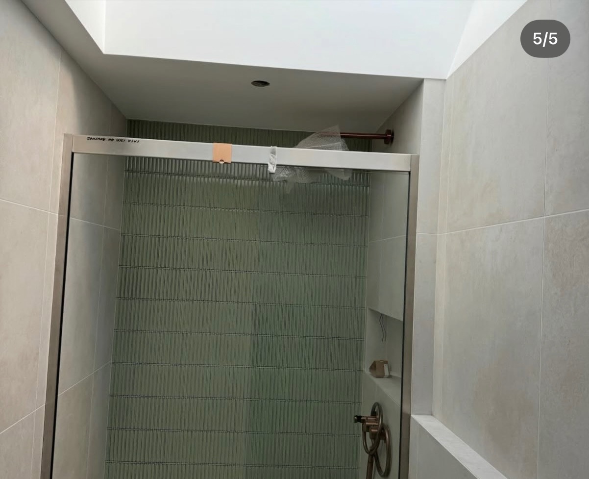 Nowoczesna kabina prysznicowa ze szklanymi drzwiami w łazience wyłożonej płytkami w odcieniach beżu. Widoczny elegancki, miedziany uchwyt i detale.