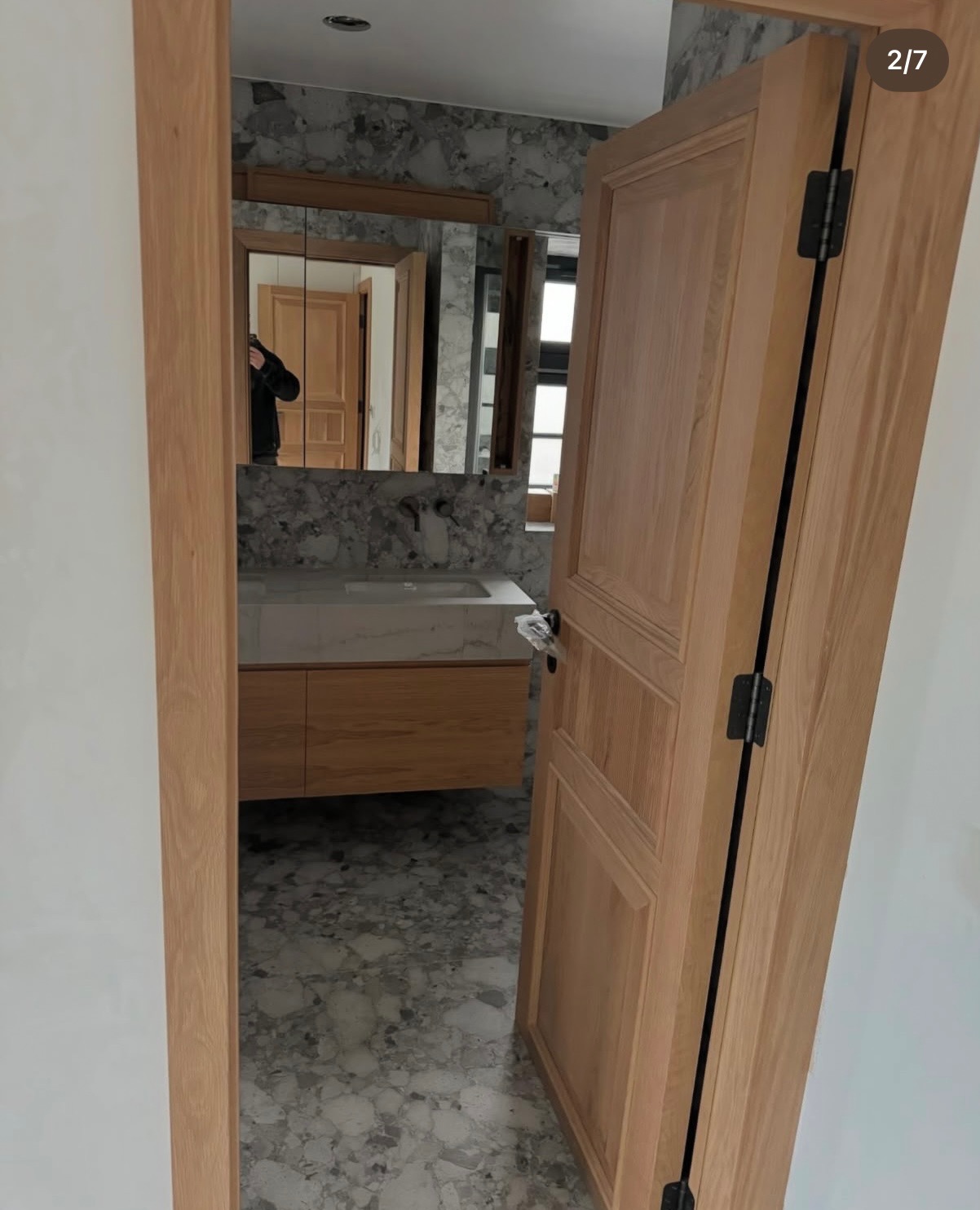 Otwarta, drewniana rama drzwi wejściowych do łazienki z marmurową podłogą i umywalką z szafką. W lustrze odbija się osoba.